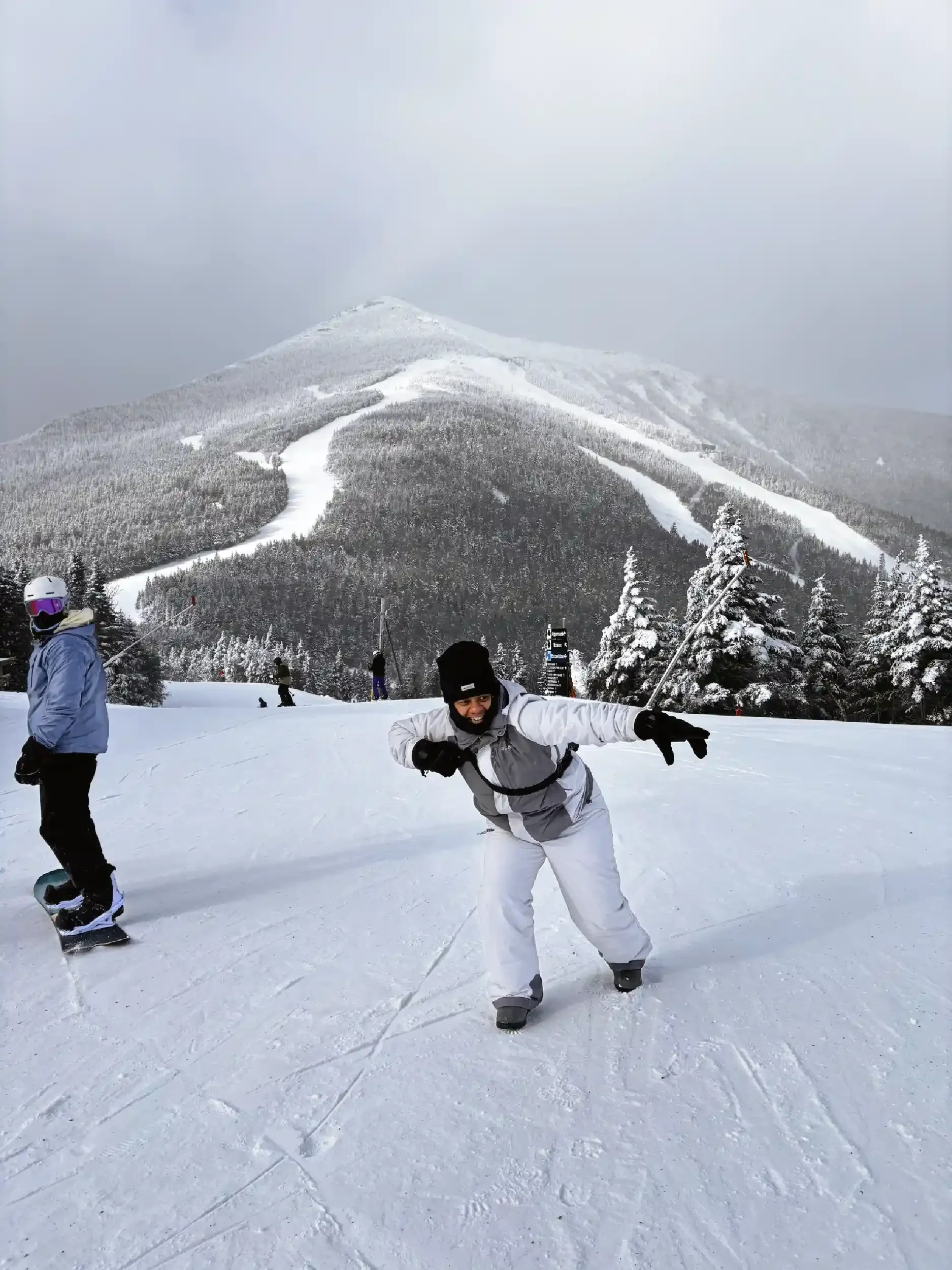 Person in white ski outfit and black gloves posing on snow-covered ski slope, mountain with ski trails and trees in the background.