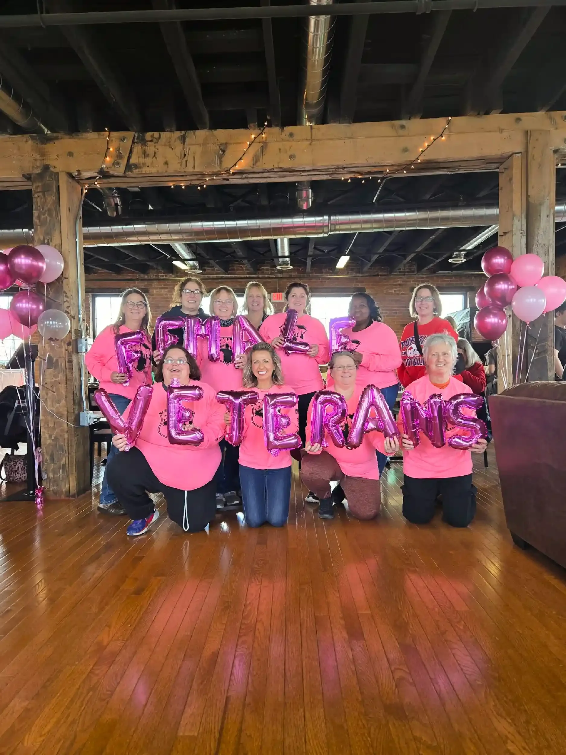 Group of women holding pink balloon letters spelling 'FAMILY VETERANS' at an indoor celebration with balloons and festive decorations.
