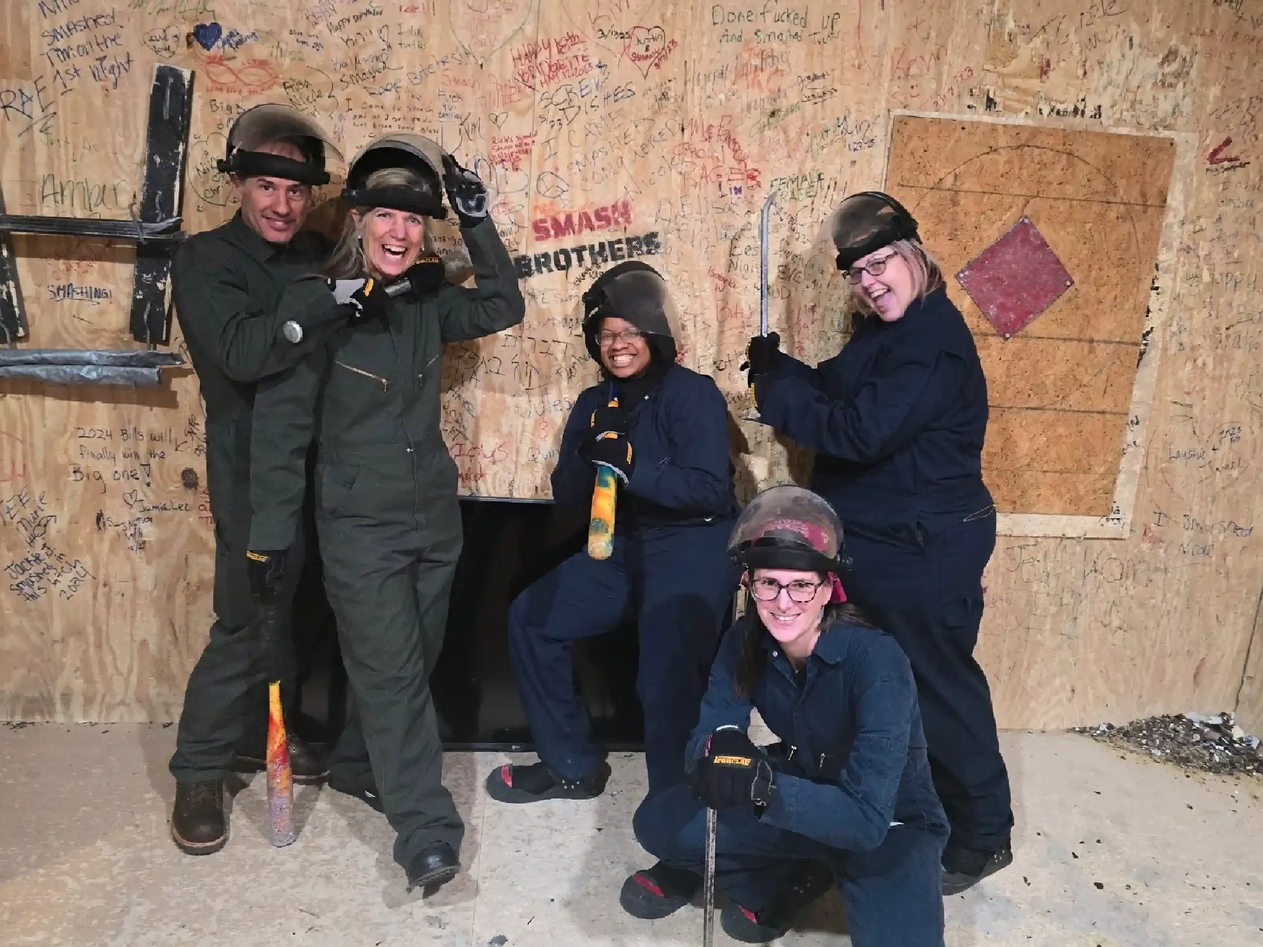 Five women in safety gear smiling and posing with tools inside a wooden-walled indoor axe-throwing area.