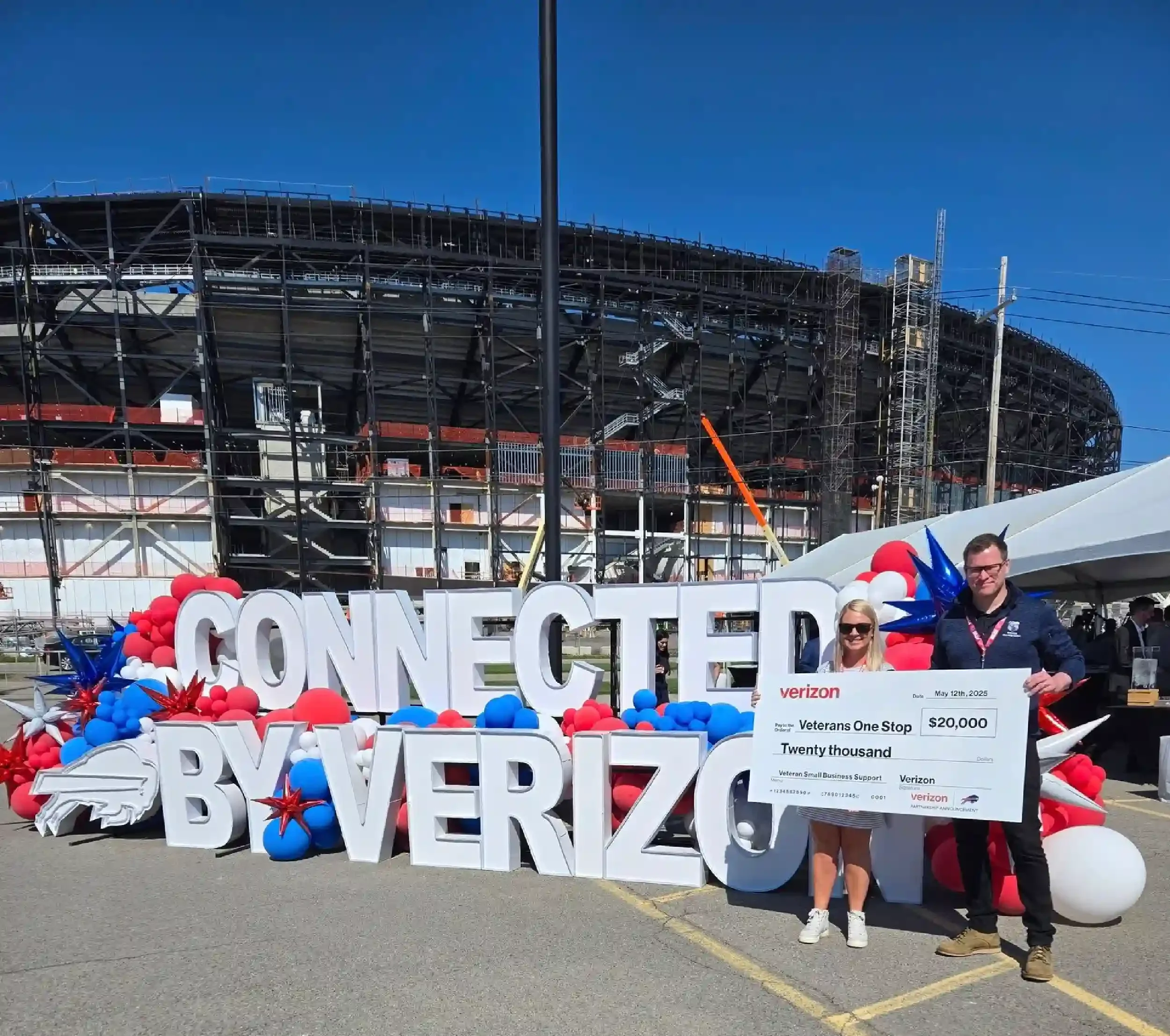 Two people standing in front of large white letters spelling 'Connect by Verizon', decorated with red, white, and blue balloons and star-shaped decorations, holding a large check for $20,000 from Verizon, outdoors at a stadium under construction with blue sky.