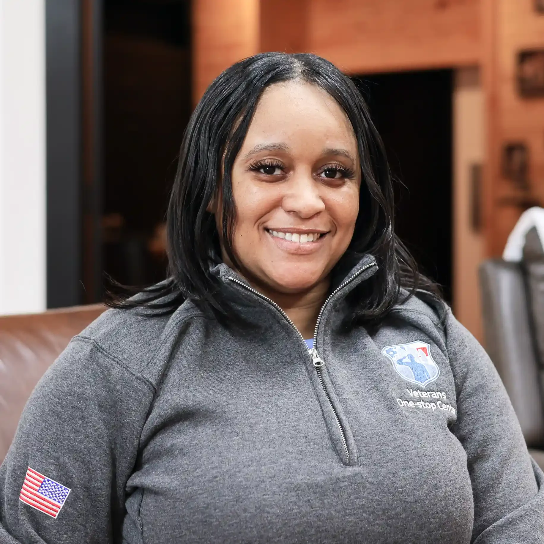 A woman with dark shoulder-length hair smiling, wearing a gray zip-up jacket with veterans' logos, sitting in a room with wood-paneled walls.
