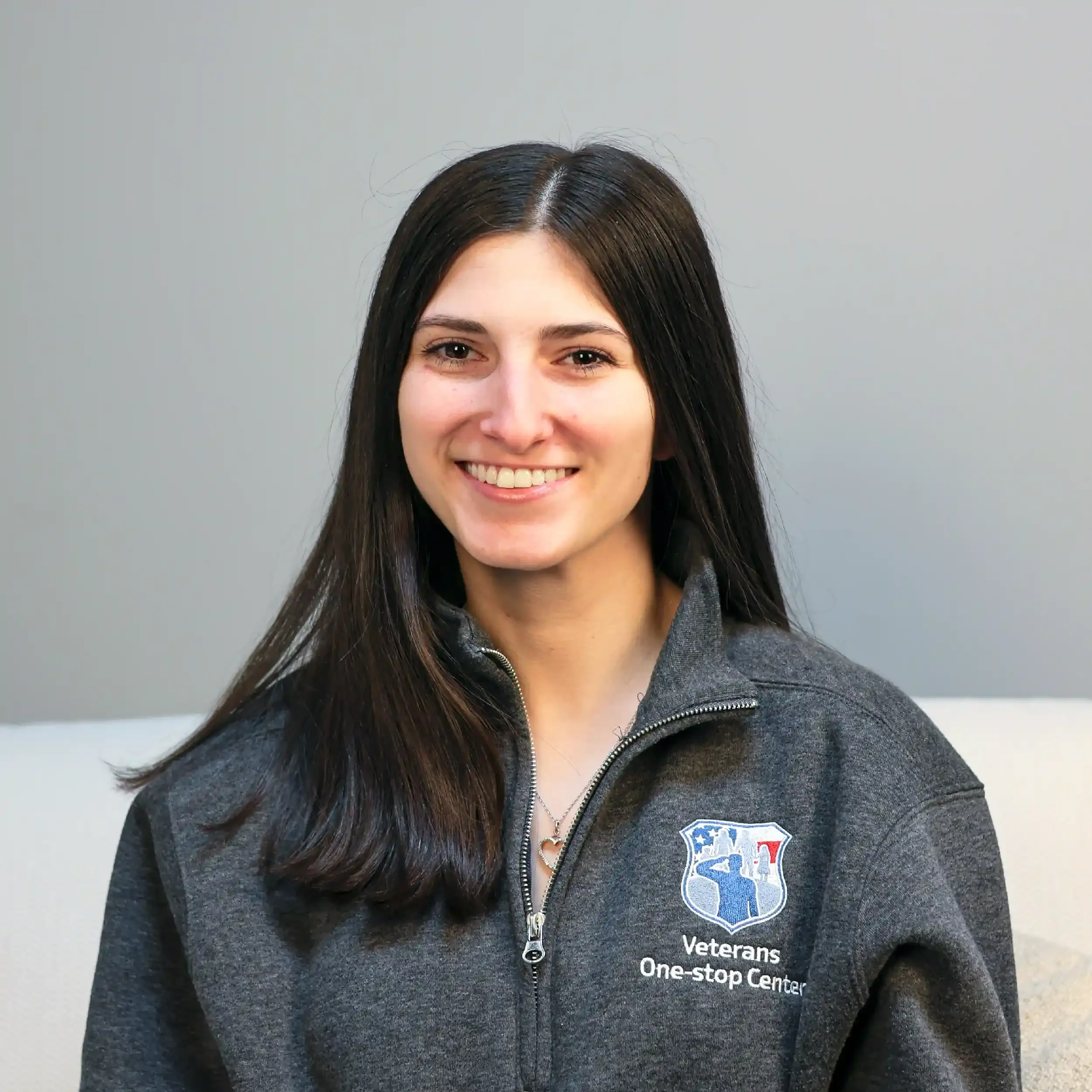 A woman with long dark hair smiling, wearing a gray zip-up jacket with a Veterans One-stop Center logo.