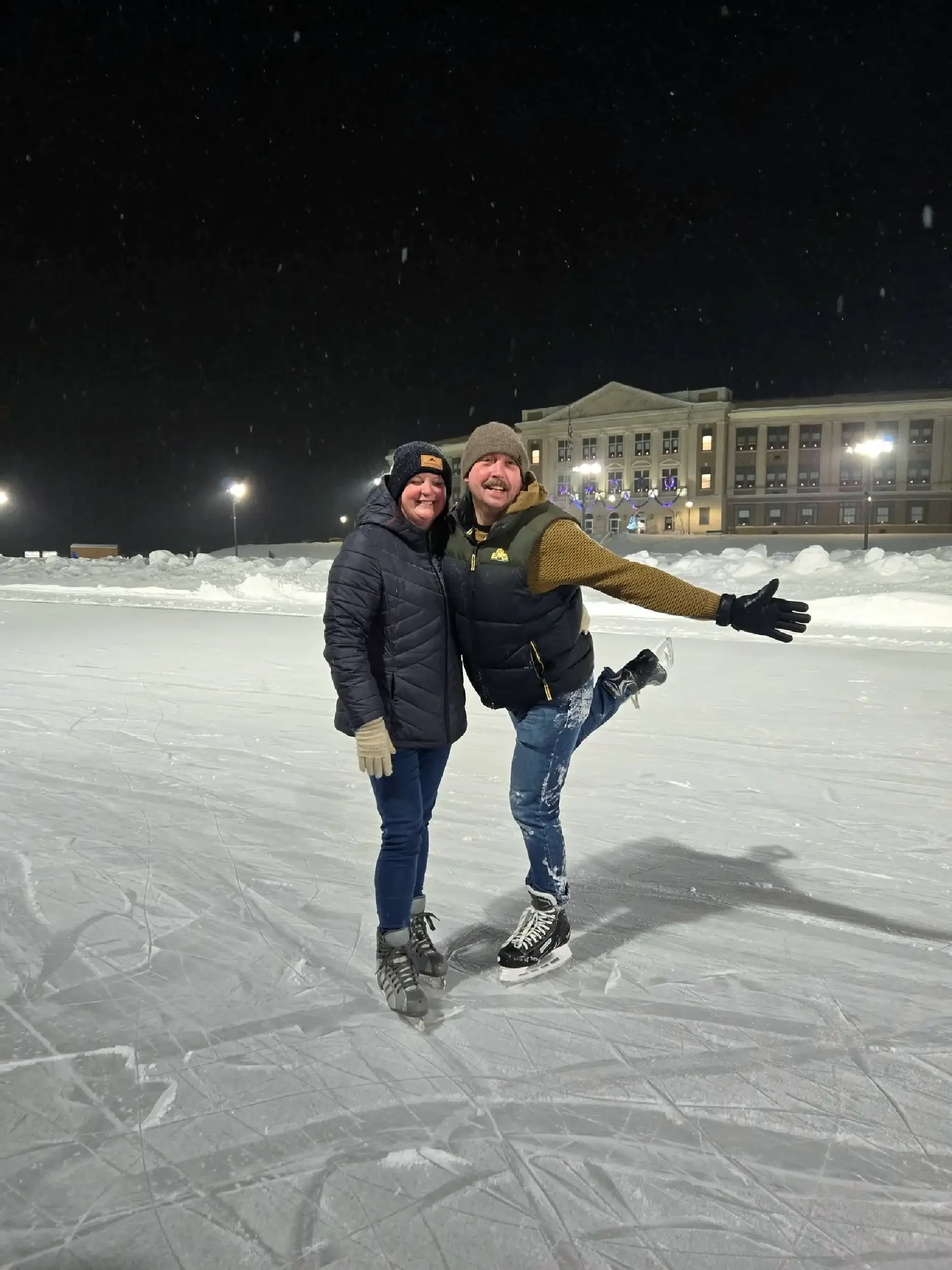 Two people ice skating on a rink at night, smiling and posing for the photo with a large building and snow-covered surroundings in the background.
