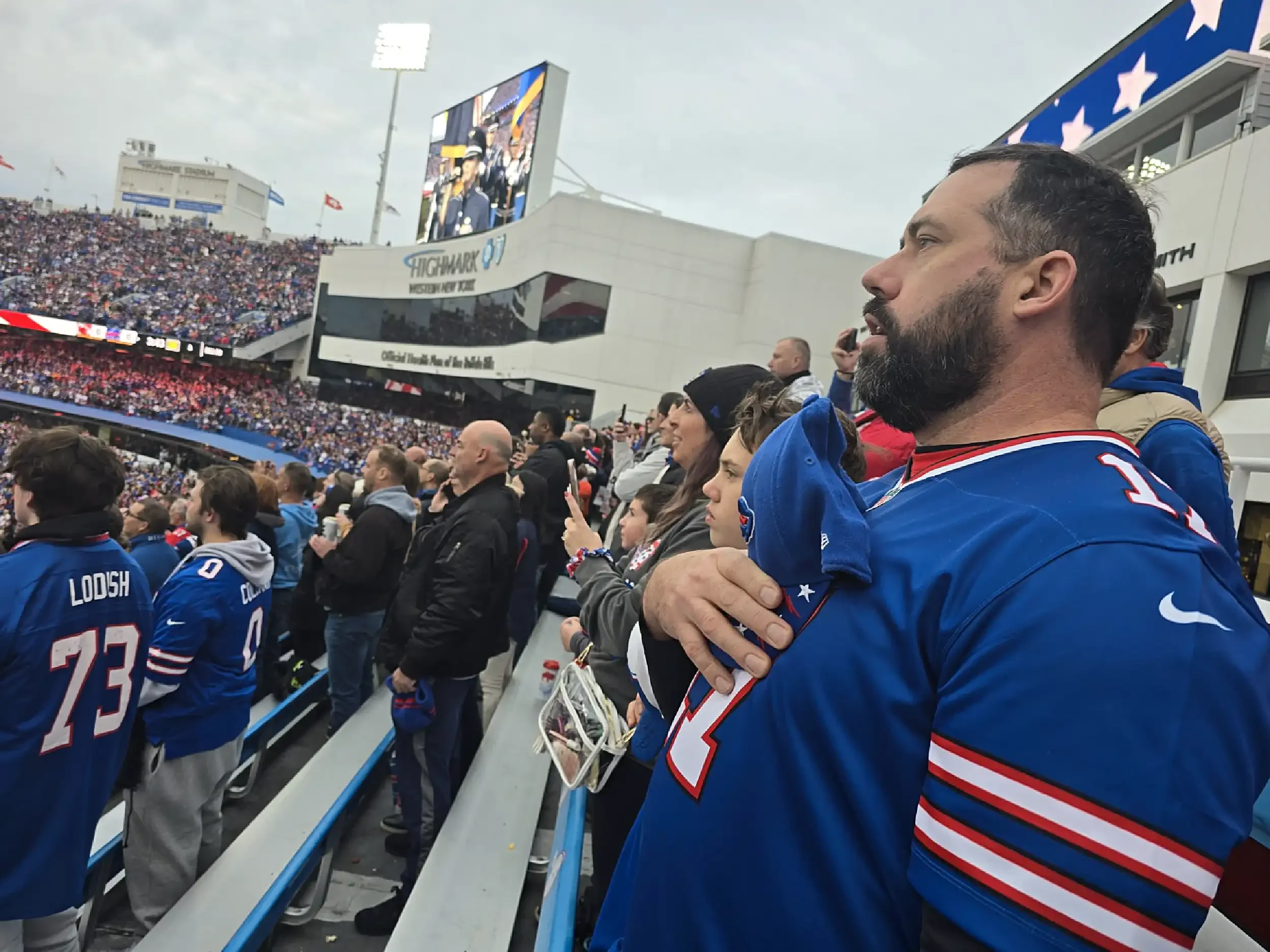 A man in a blue football jersey holding a child with a similar jersey at a stadium, with other spectators and football players watching a game on the field.