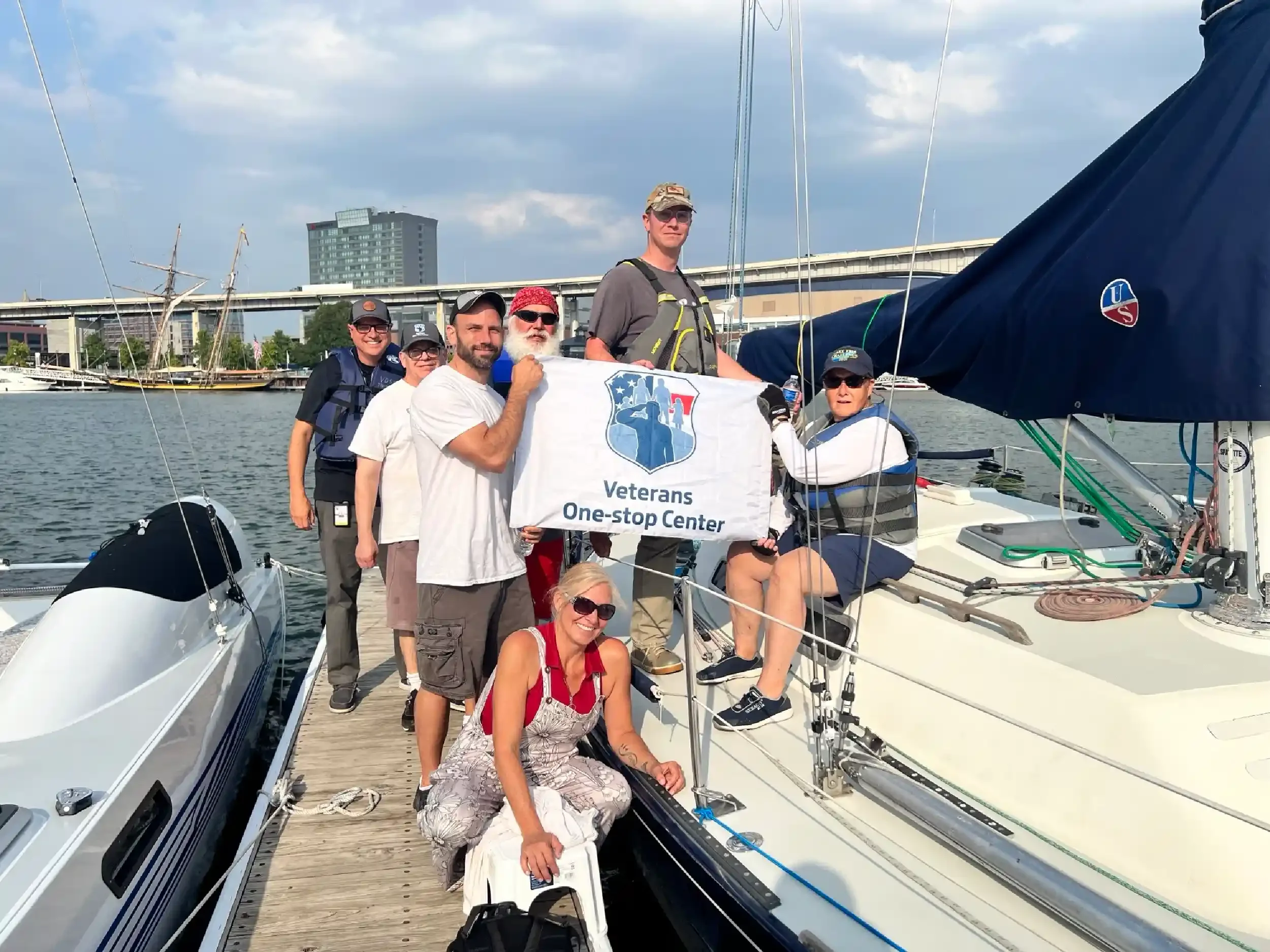 Group of people on a boat dock holding a Veterans One-stop Center banner, smiling during a sunny day with water and boats in the background.
