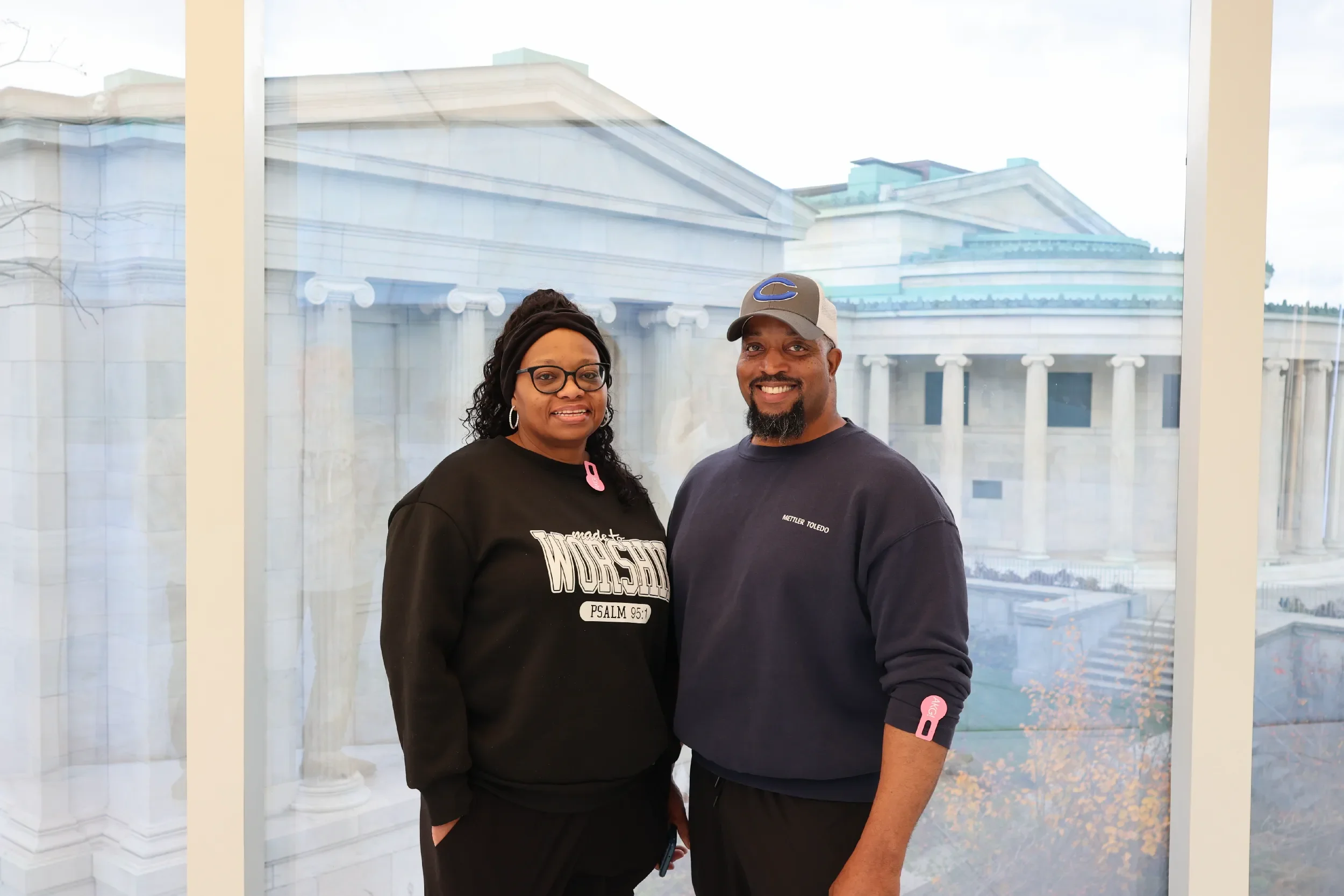 A woman and man smiling indoors in front of a large window with a view of the Capitol building.