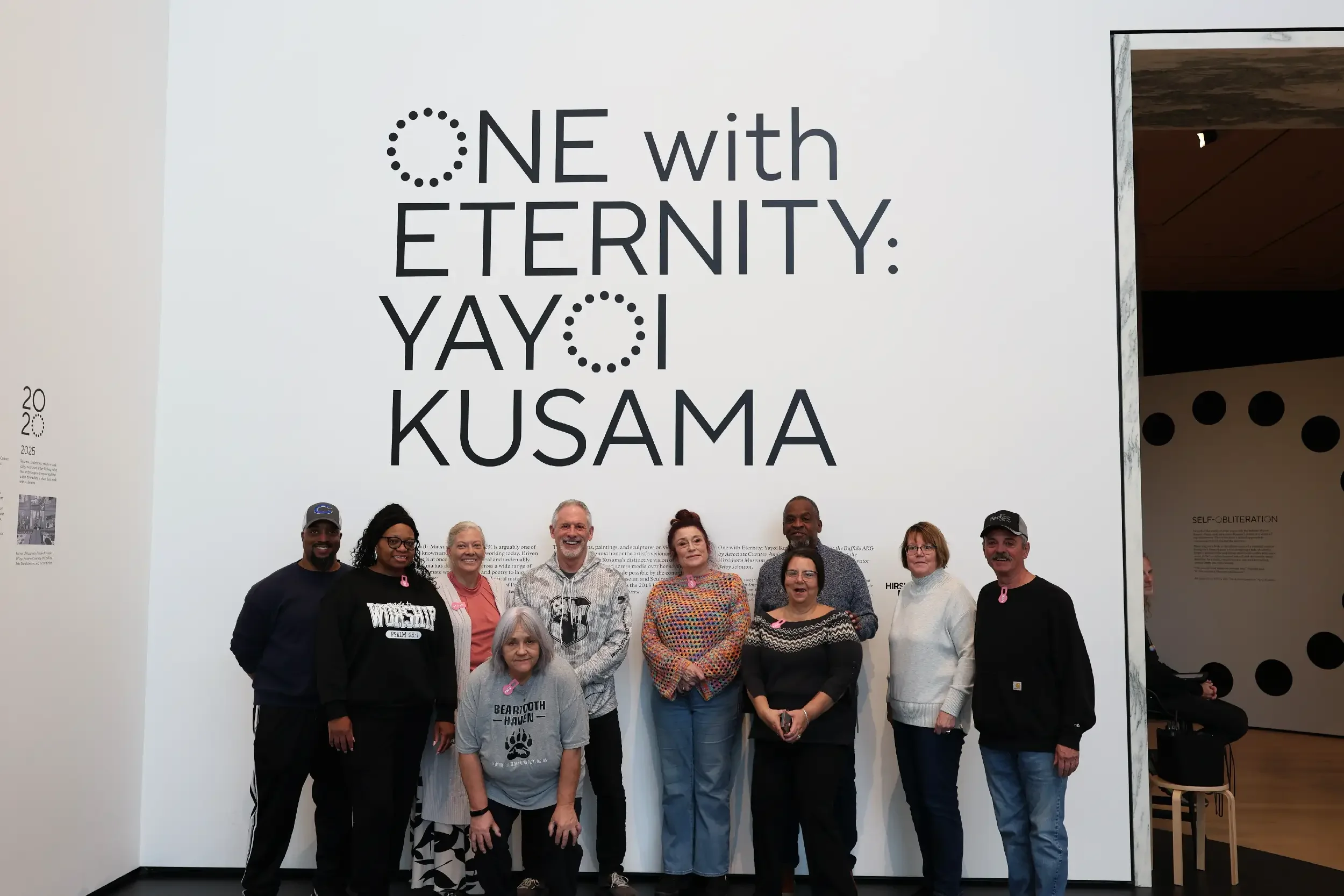 A group of ten diverse people standing in front of a large white wall with black text that reads 'ONE with ETERNITY: YAY I KUSAMA'.
