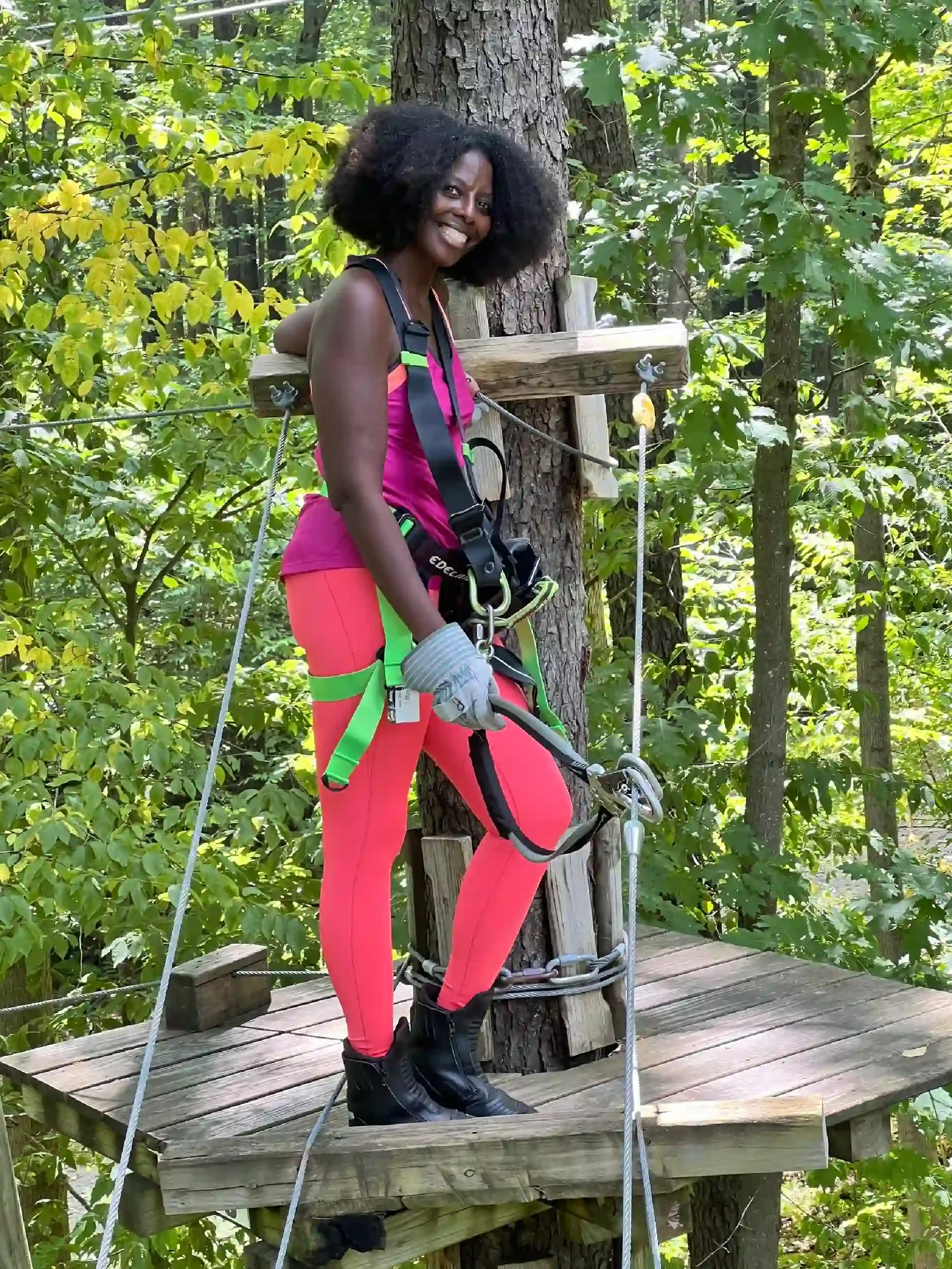 A woman with dark curly hair and dark skin standing on a wooden platform in a forested area, wearing climbing harness, gloves, black boots, and pink and green activewear, smiling at the camera.