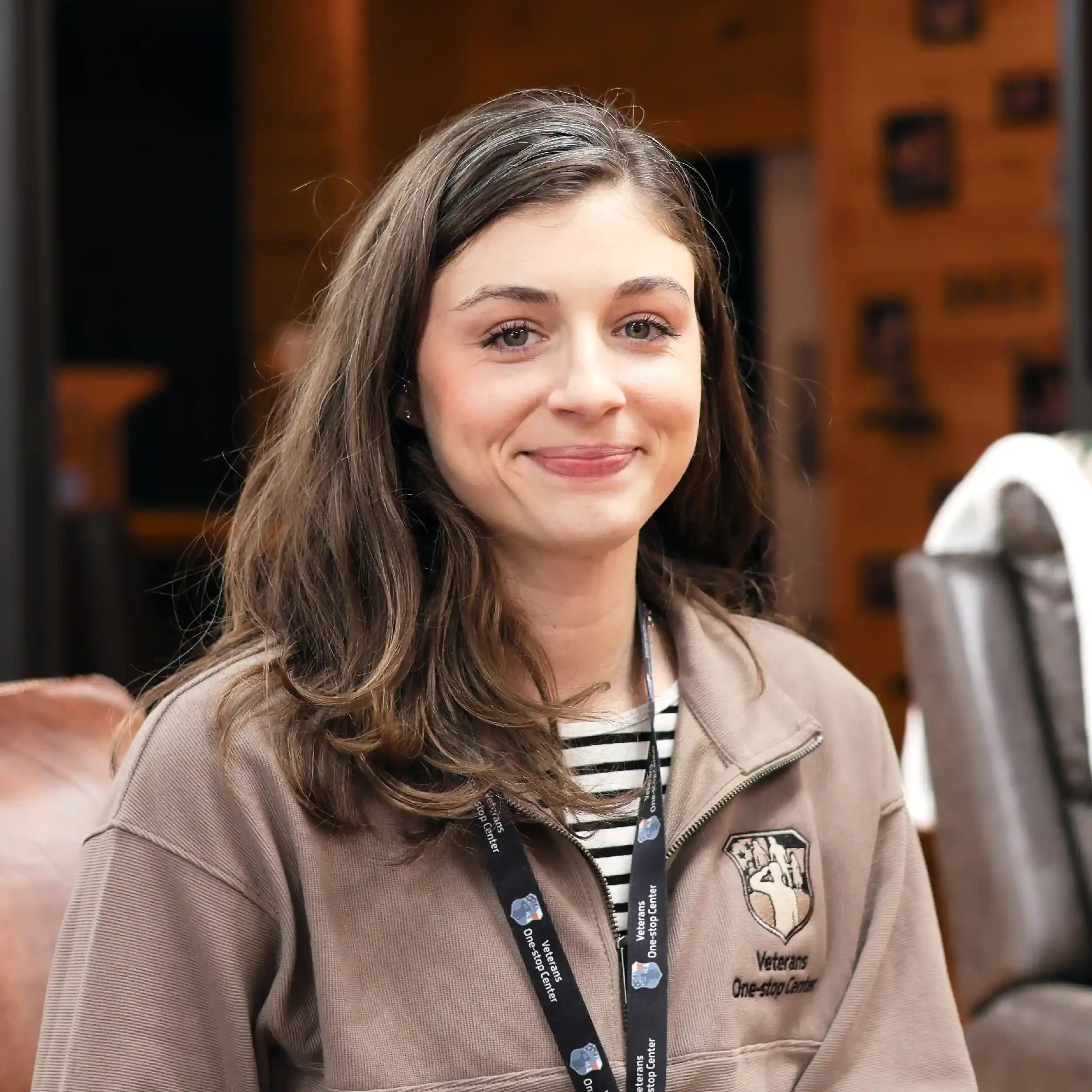 Young woman with brown hair smiling, wearing a beige jacket with a Veterans One-stop Center logo, and a black lanyard, sitting inside a wood-paneled room.