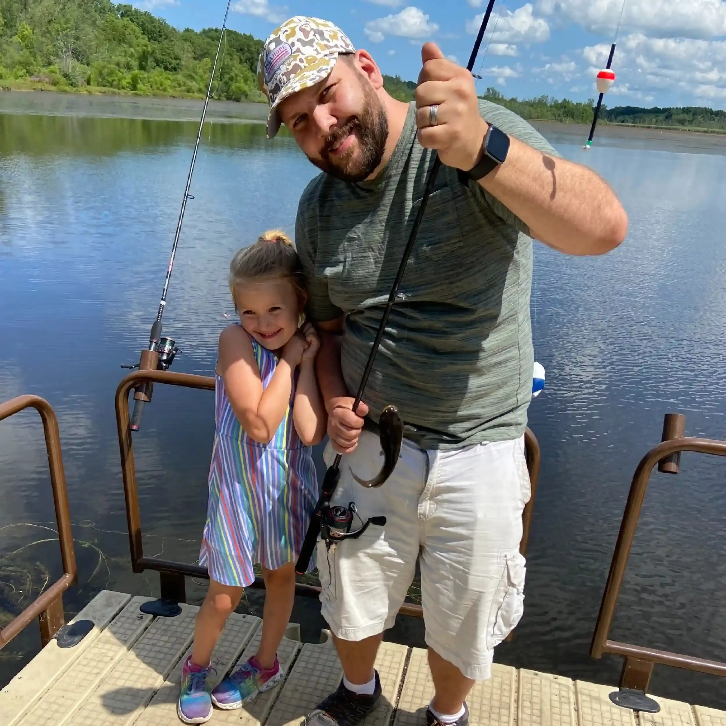 A man and a young girl on a dock by a lake, fishing and smiling at the camera, with fishing rods in their hands and water and trees in the background.