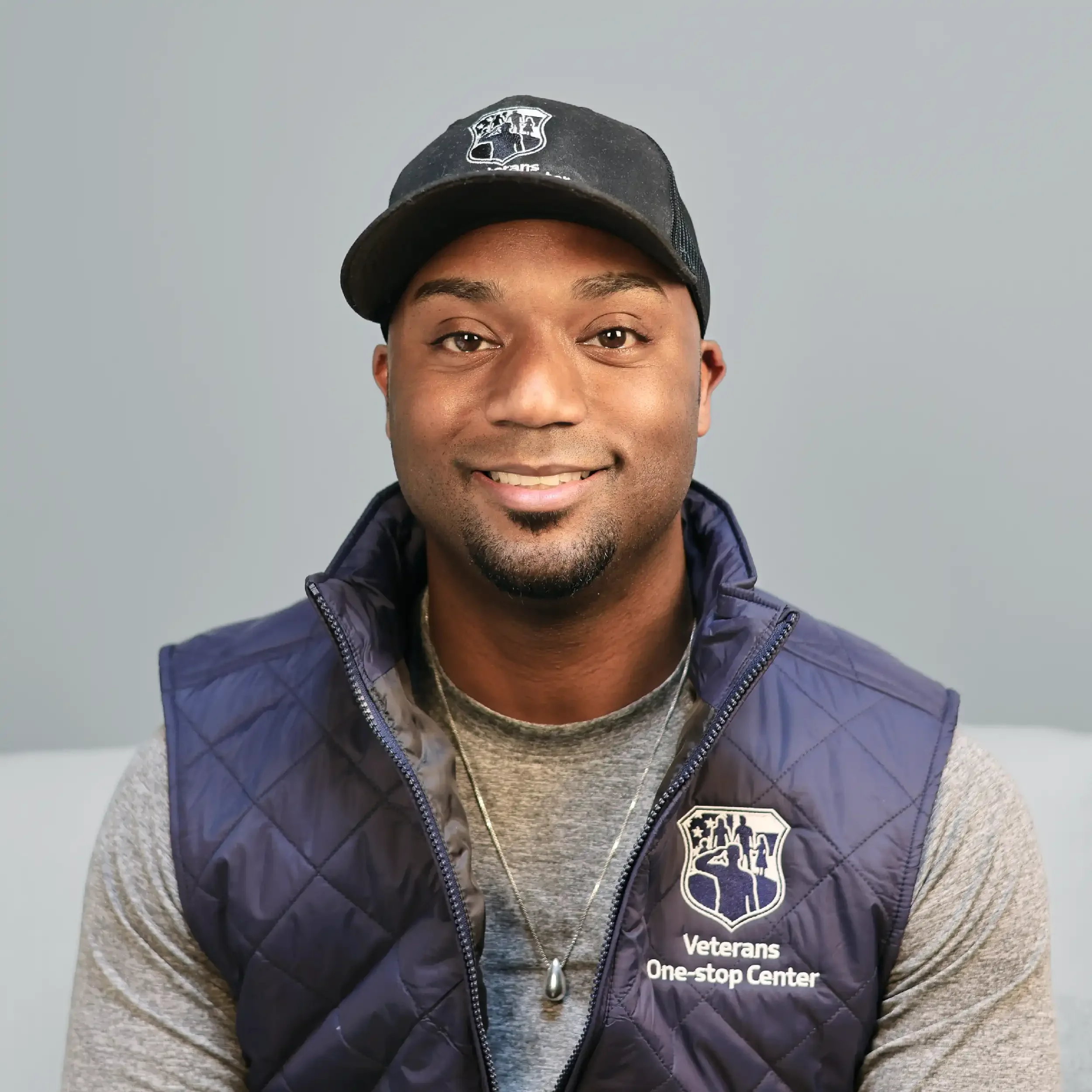 A man wearing a black cap, gray shirt, and navy vest with a Veteran's One-stop Center logo, smiling against a plain gray background.