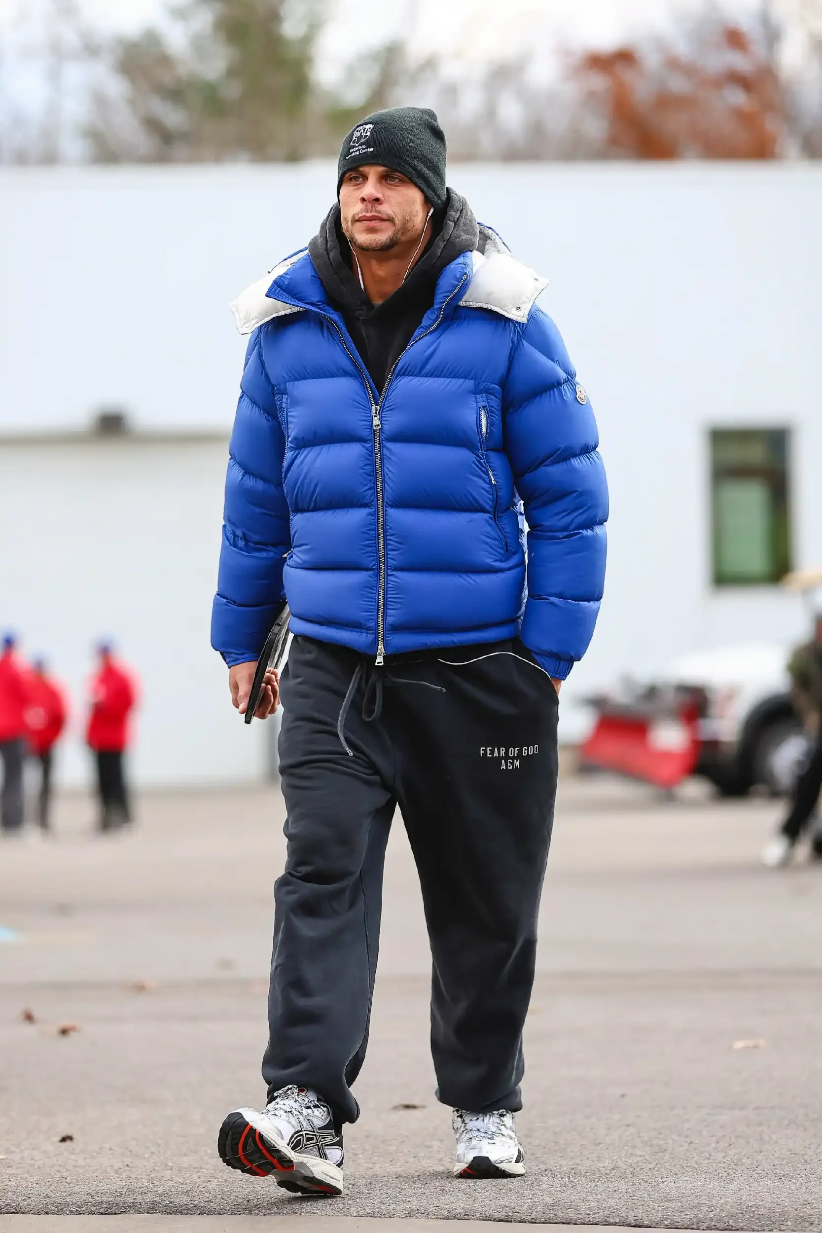 A man in a blue puffer jacket, black beanie, black sweatpants, and white sneakers walks outdoors, with a few people and cars in the background.