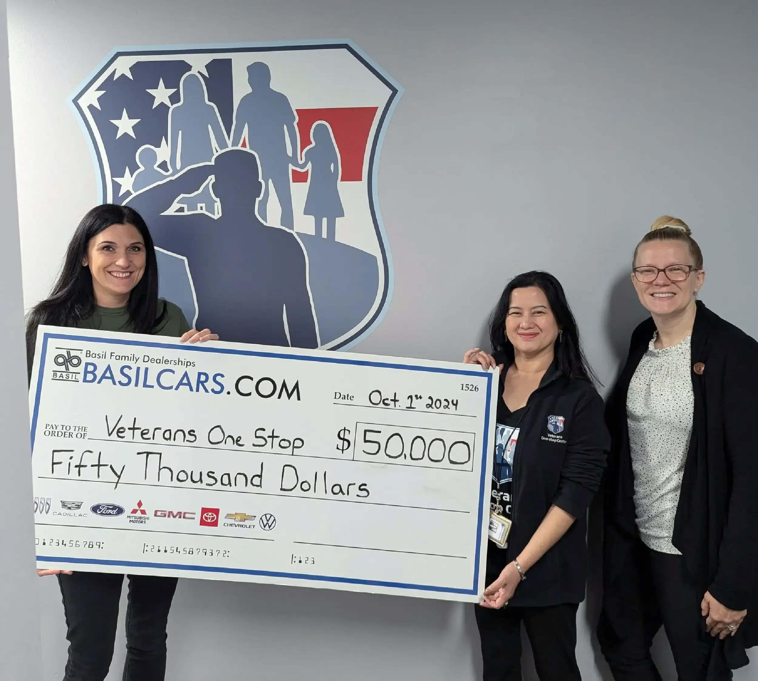 Three women holding a large check from Basil Family Dealerships for fifty thousand dollars, made out to Veterans One Stop, date October 1, 2024, standing in front of a wall with a family-themed logo.