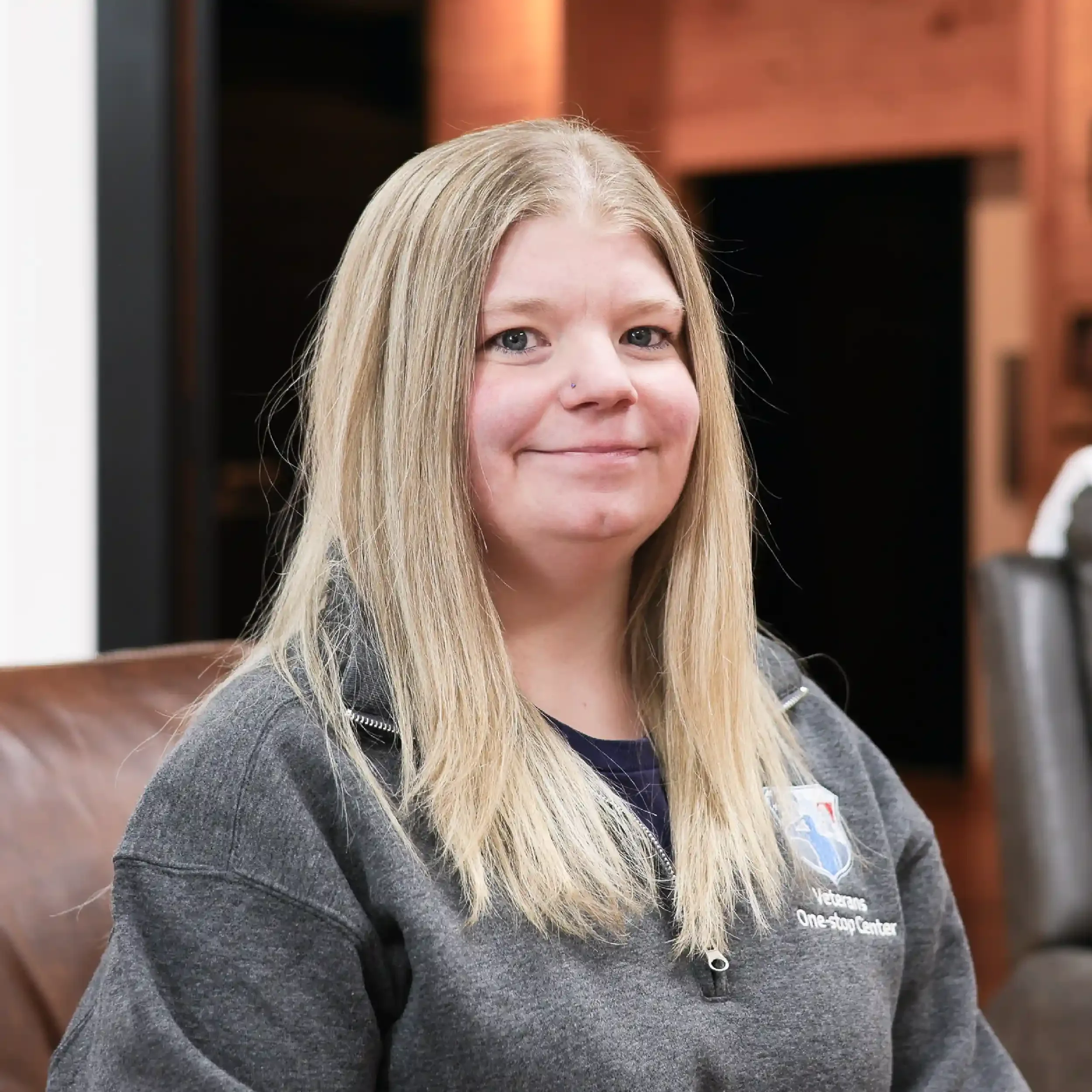 A woman with long blonde hair and blue eyes, wearing a gray quarter zip jacket with a logo and text on it, sitting in an indoor setting with wooden paneling and chairs in the background.