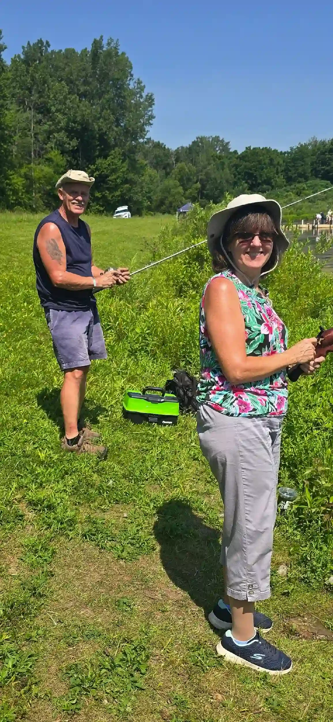 Two women standing outdoors on a sunny day, fishing in a grassy area with trees in the background.