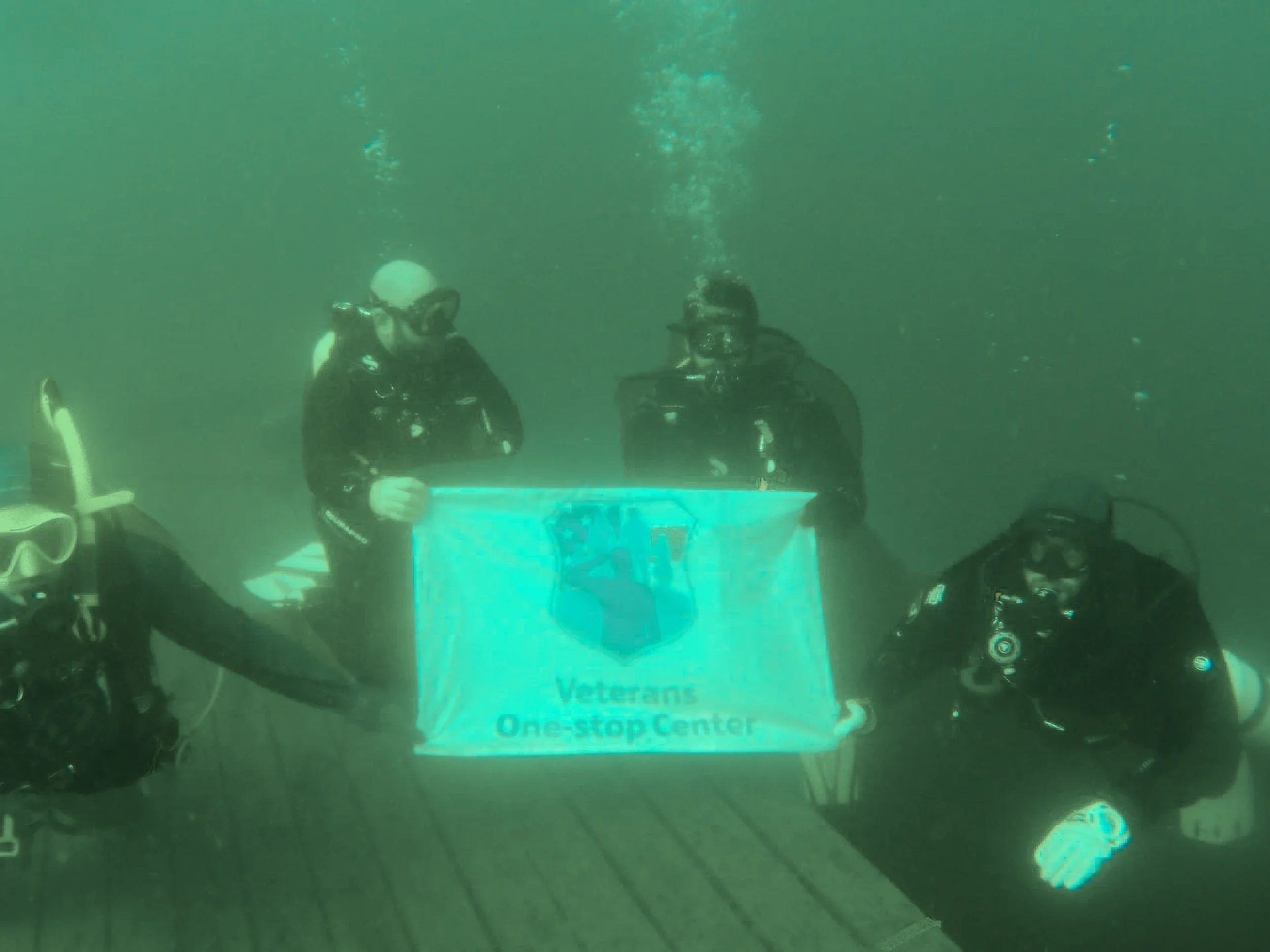 Three scuba divers underwater holding a sign that reads 'Veterans One-Stop Center.'