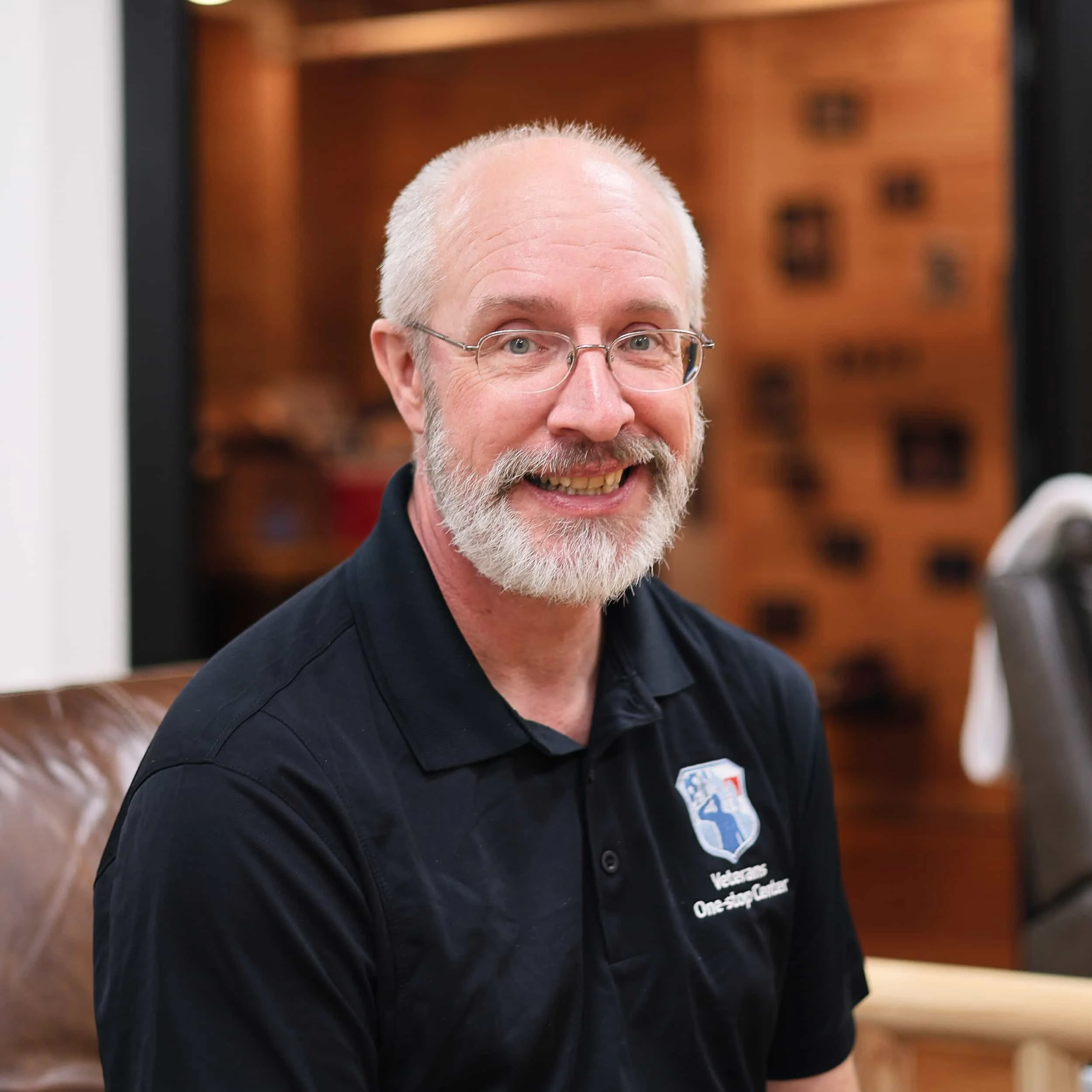 A smiling older man with a beard and glasses, wearing a black polo shirt with a logo, seated in an indoor setting with wooden walls in the background.
