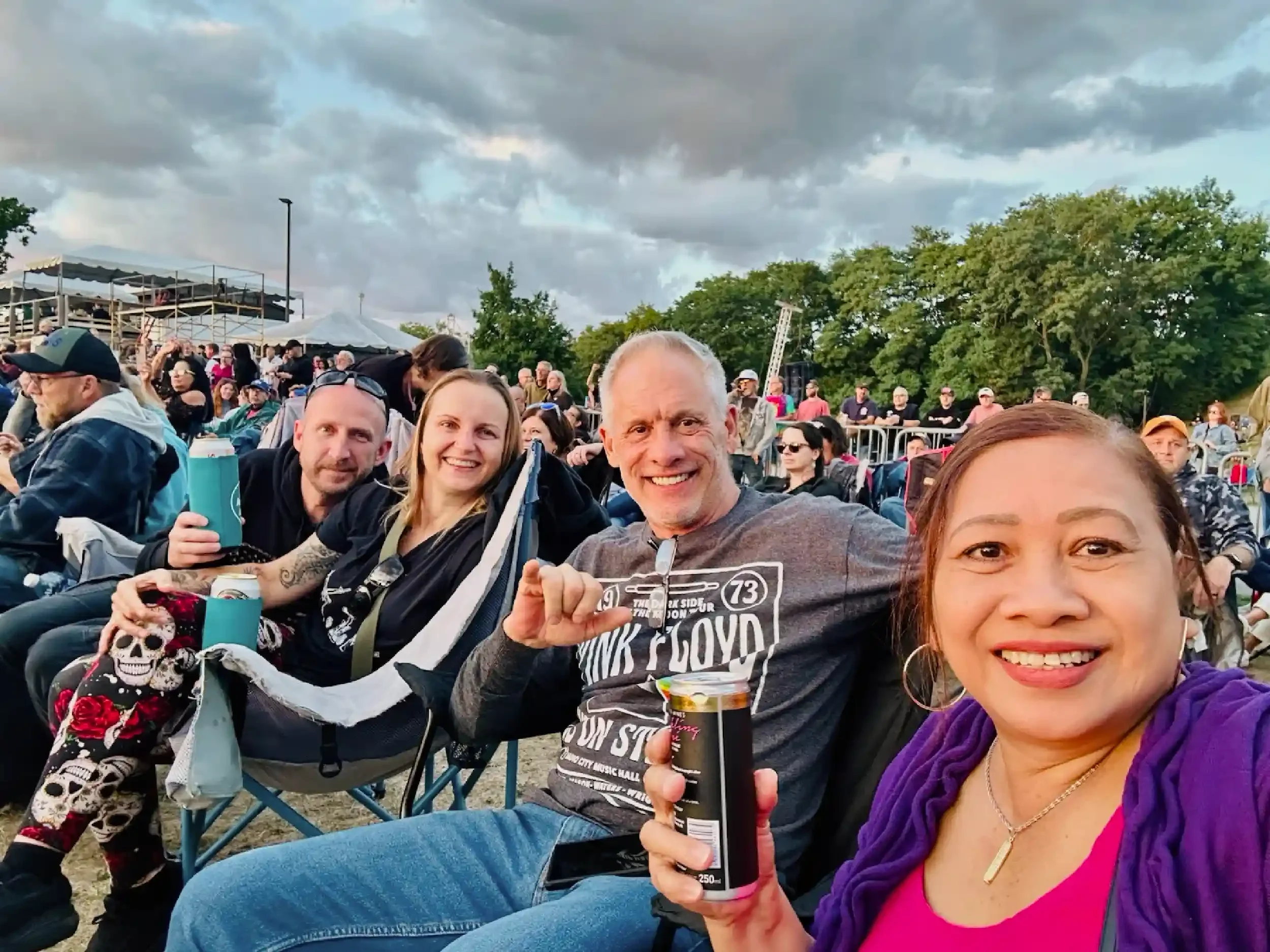 Group of five people sitting outdoors at a concert, smiling and enjoying drinks, with a crowd and trees in the background under a cloudy sky.