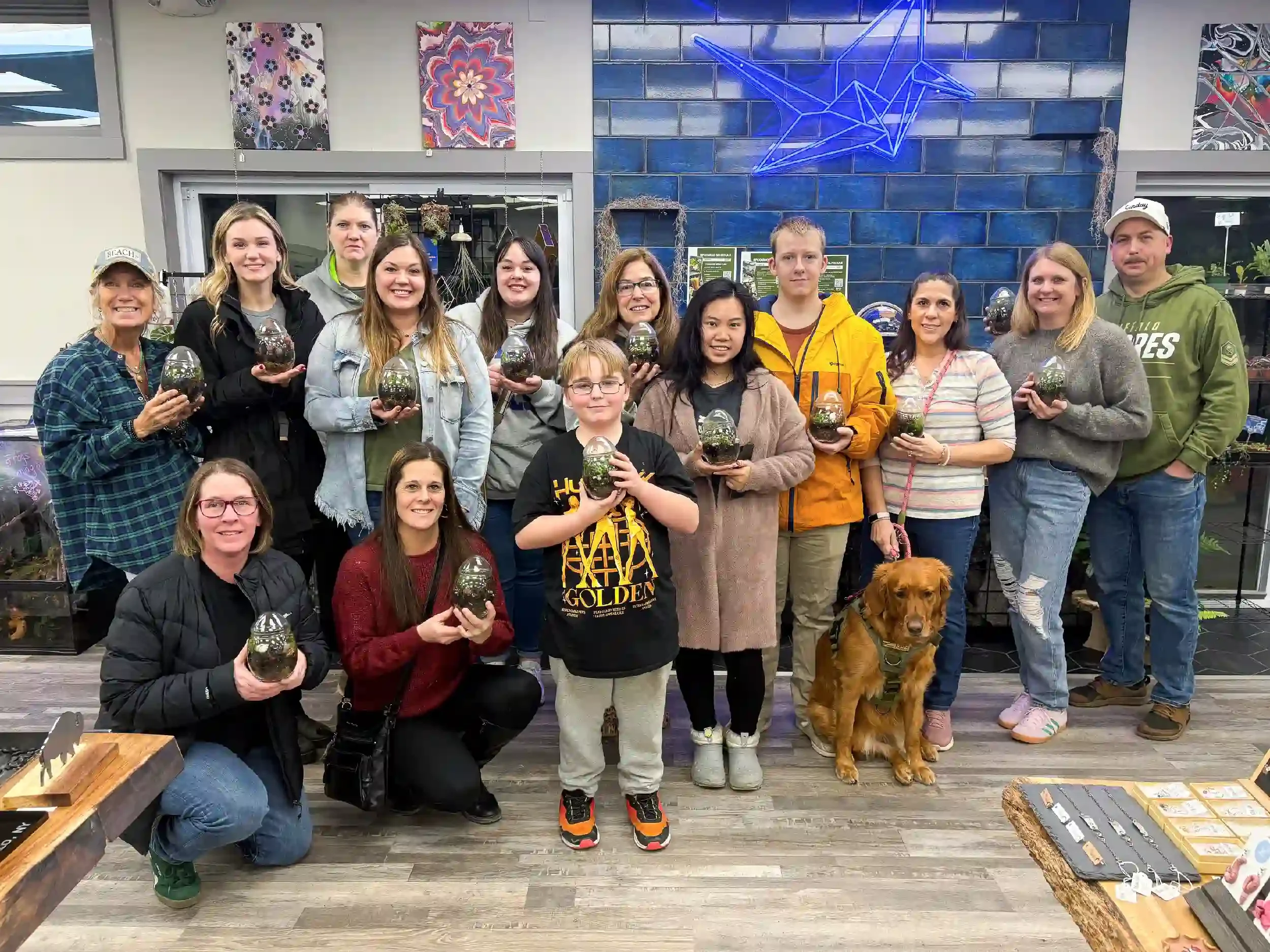 Group of people gathered in a store, holding glass terrariums with plants inside, alongside a golden retriever dog.