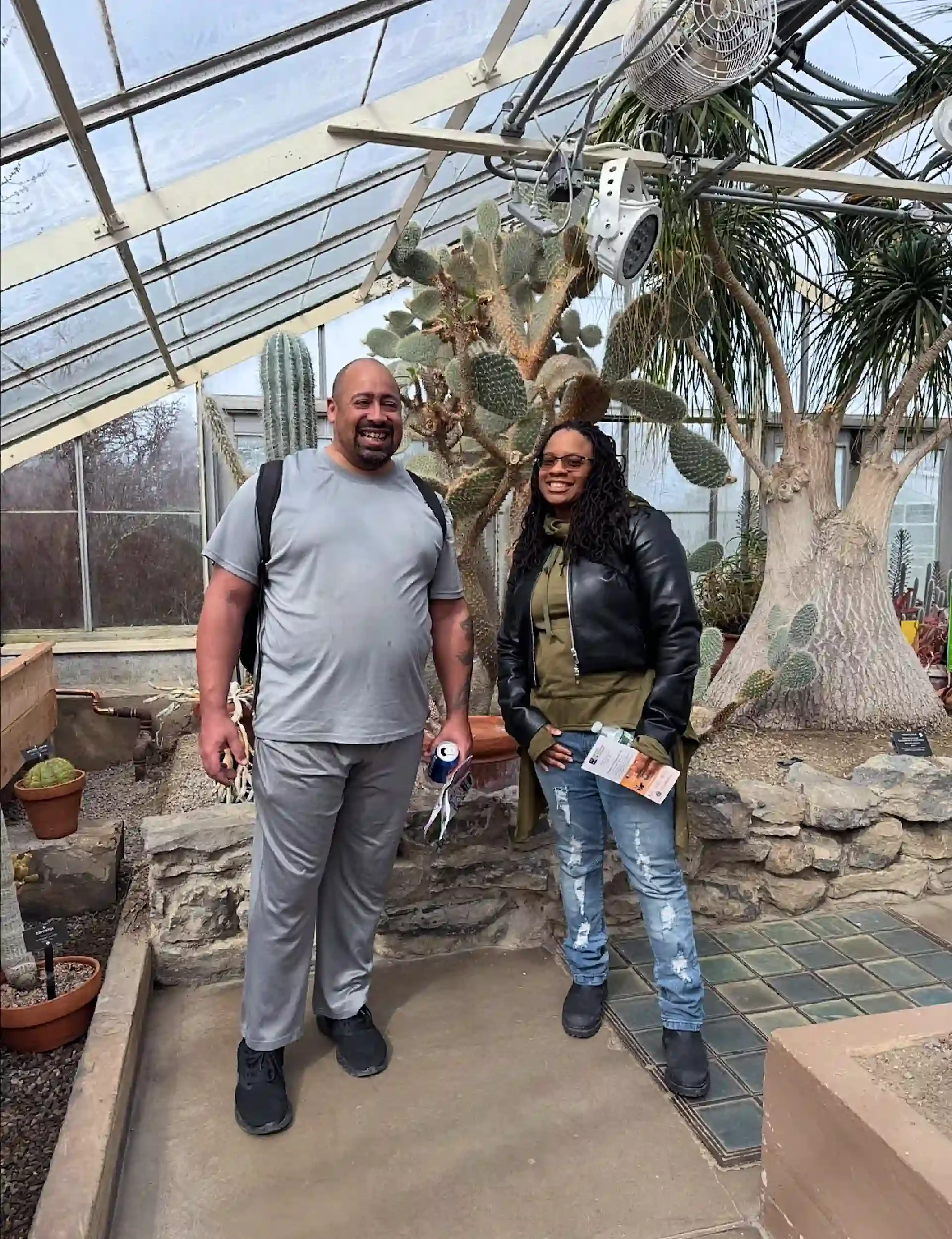 Two people smiling inside a greenhouse surrounded by large cacti and succulents.