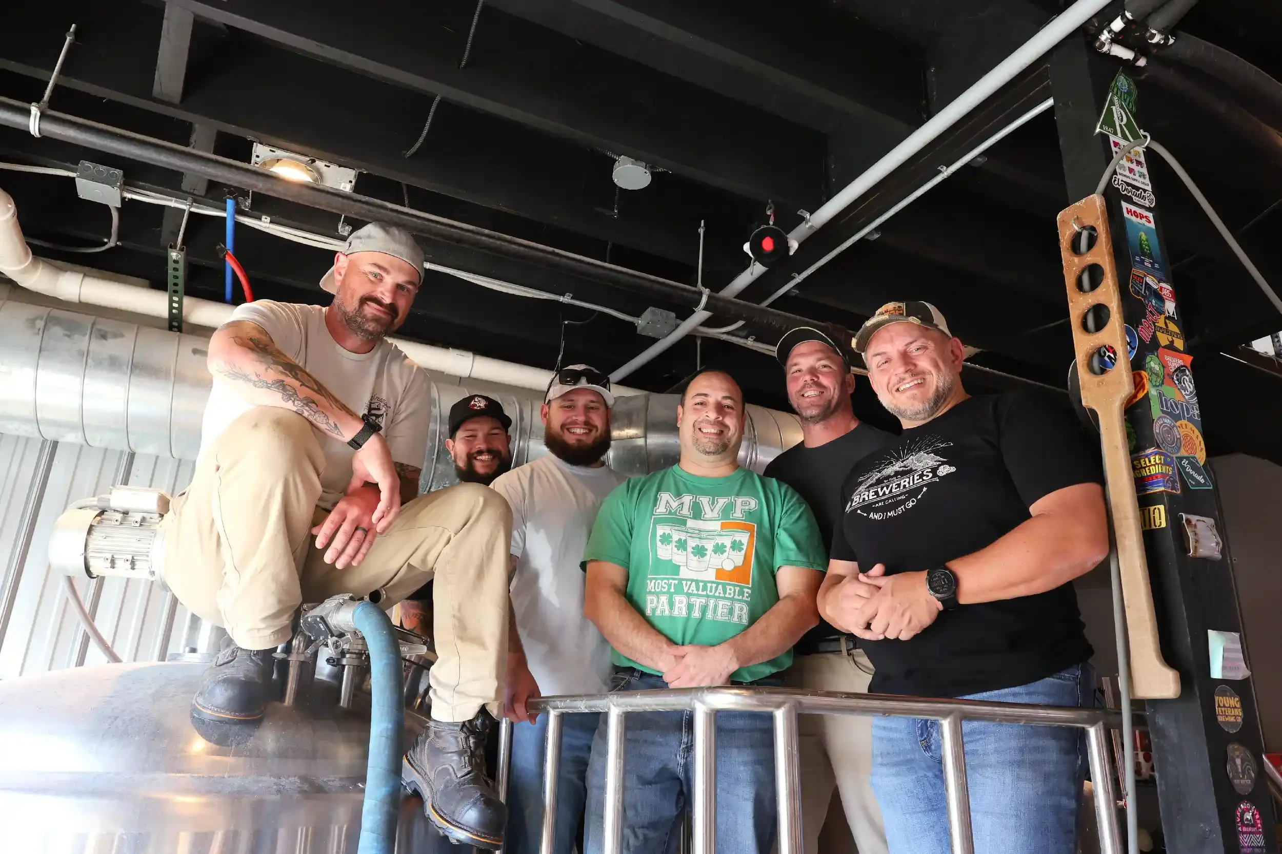 Group of six smiling men in casual clothing, standing together in an industrial setting, some wearing hats, with brewery equipment and stickers visible in the background.