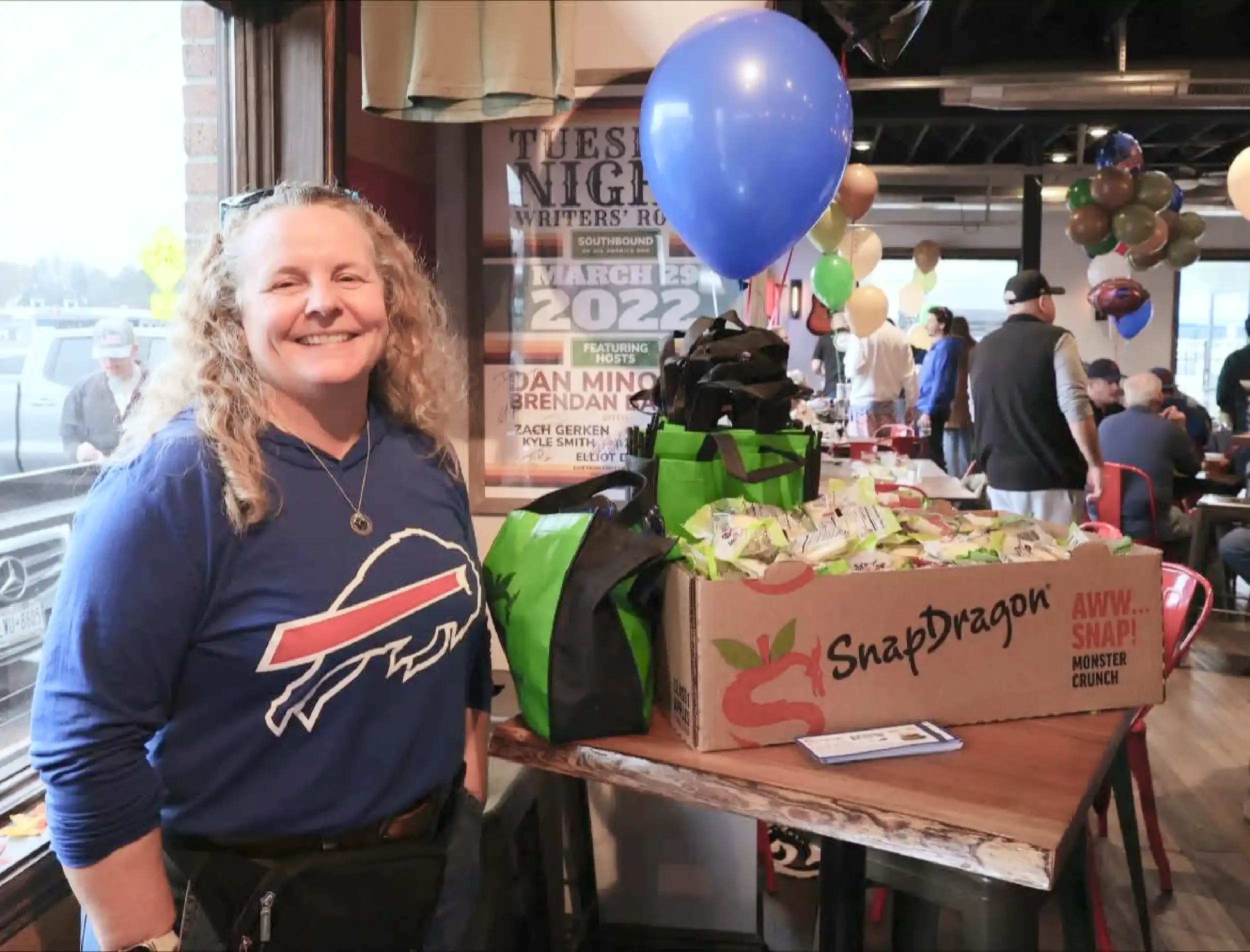 A woman with blonde curly hair smiling at a gathering indoors. She is wearing a blue shirt with a sports team logo and a necklace. Behind her and to the right, there is a table with a large box labeled 'Snapdragon', filled with snacks, and decorated with balloons. The background shows other people milling around at a social event with balloons and posters on the walls.