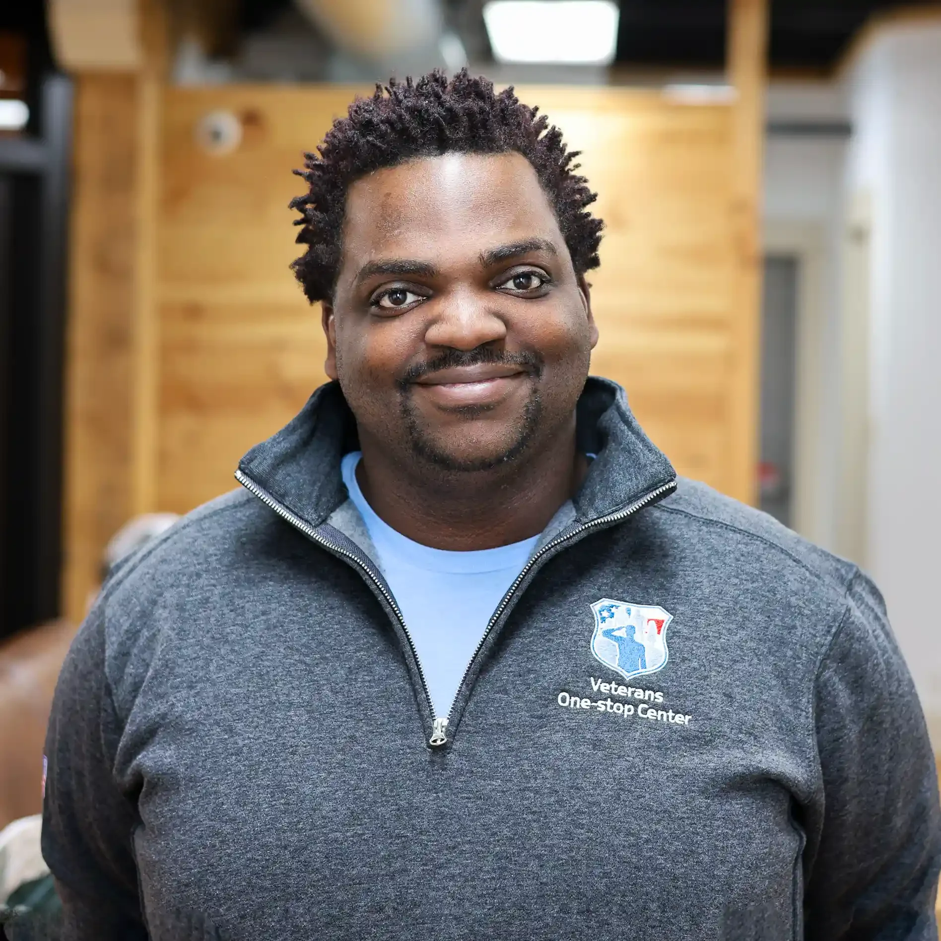 A smiling man with short curly hair wearing a gray jacket with a 'Veterans One-stop Center' logo on it, indoors with wooden paneling in the background.