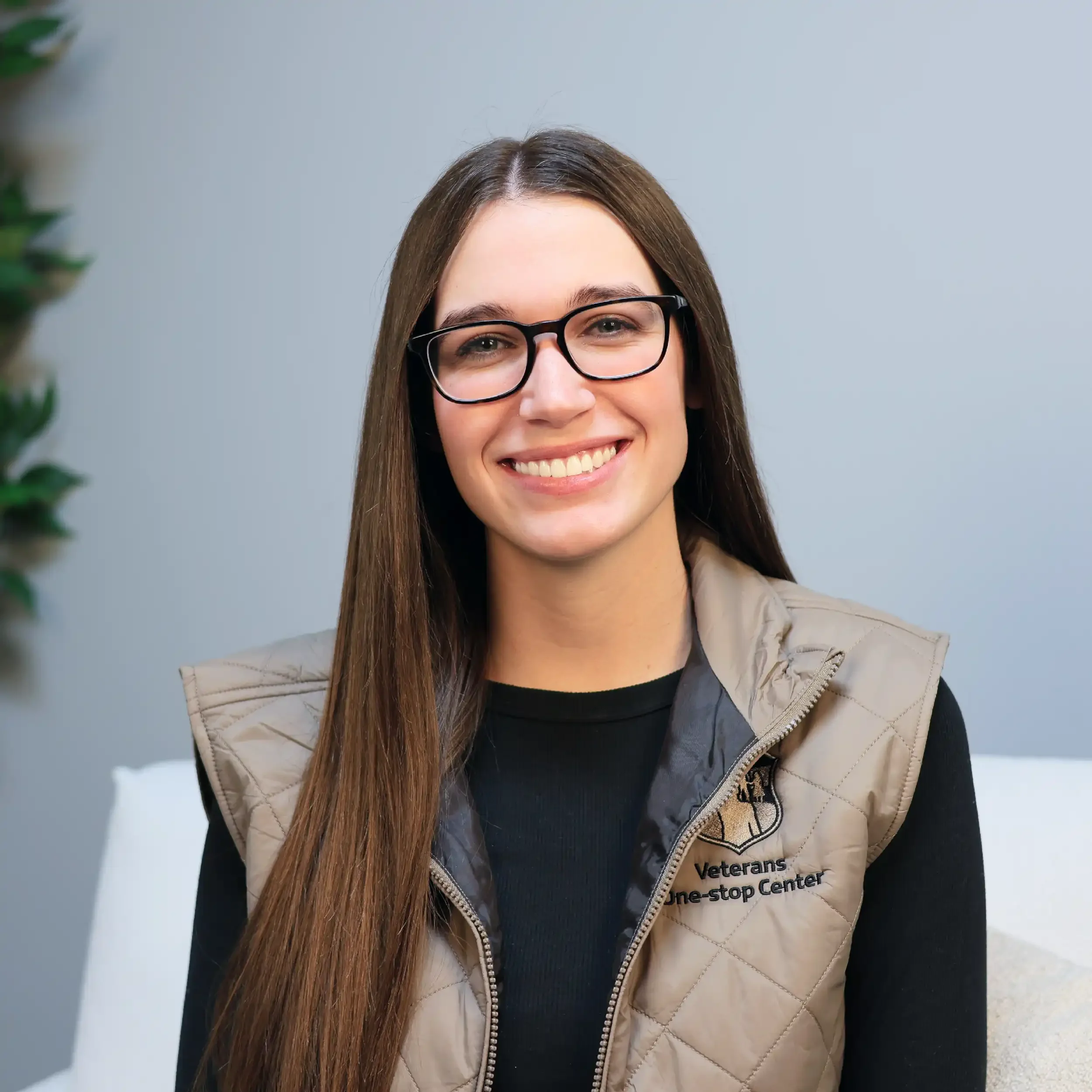 A young woman with long brown hair, glasses, and a beige vest smiling at the camera, sitting in a well-lit indoor setting.