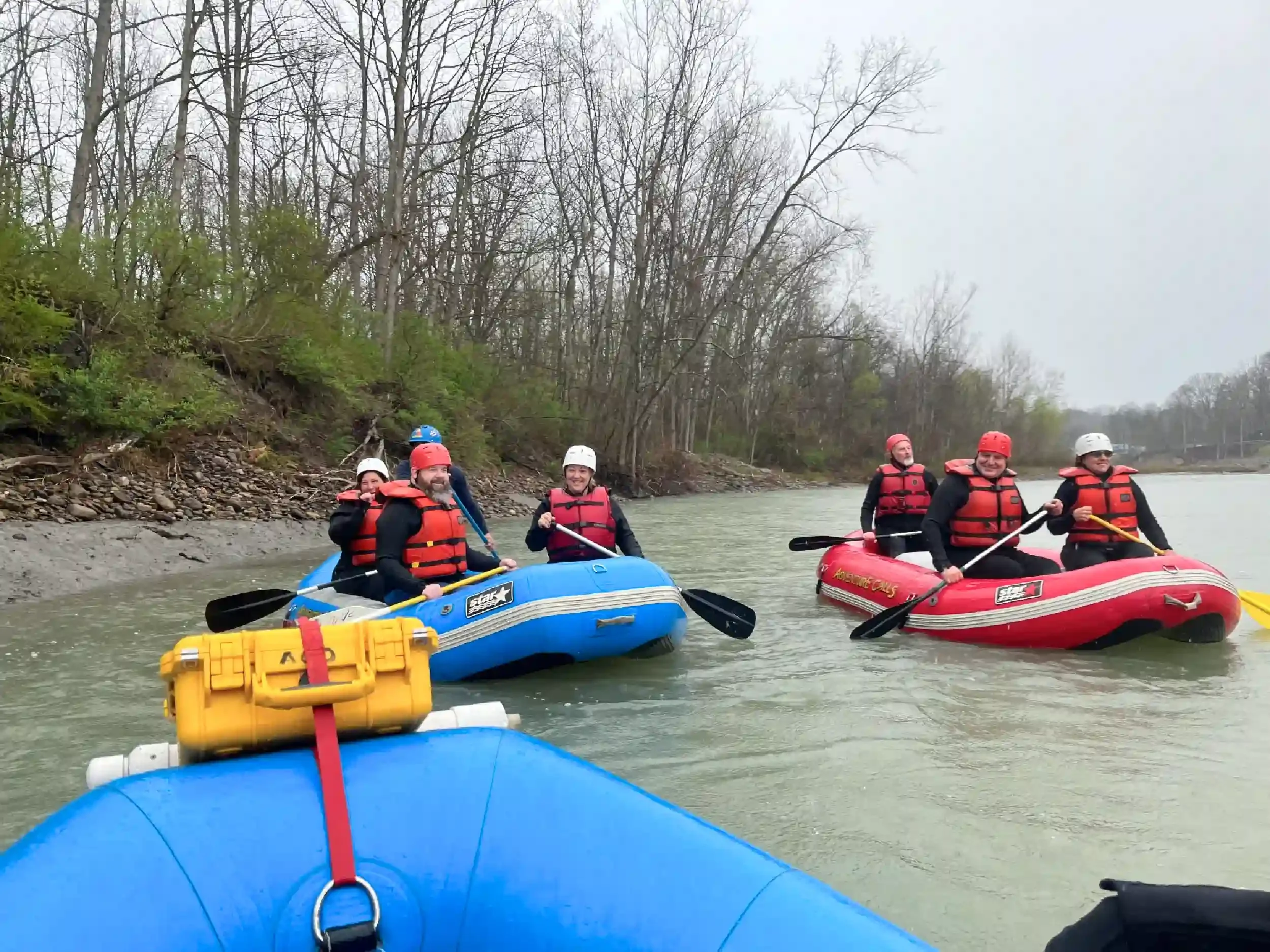 Group of six people rafting on a river, wearing life jackets and helmets, with trees along the riverbank in the background.