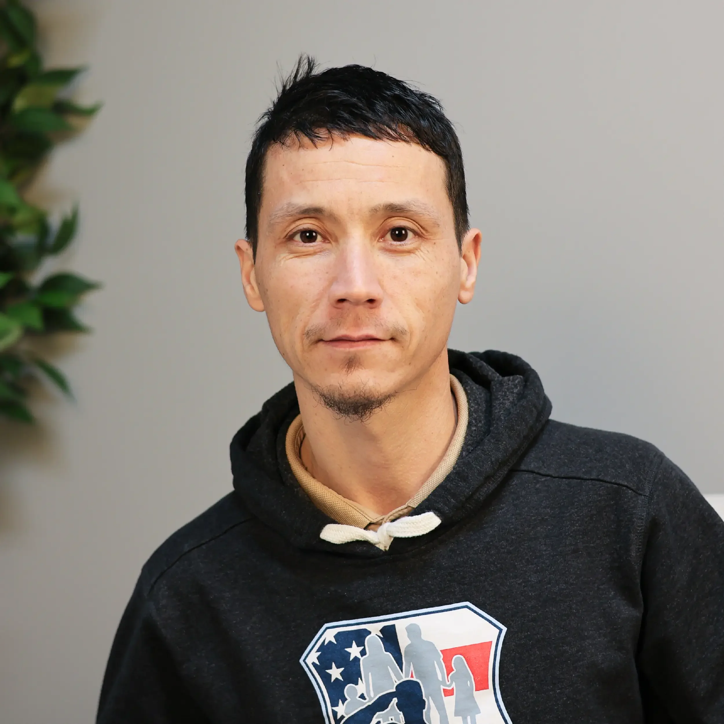 A man with short black hair and a goatee, wearing a dark jacket over a beige collared shirt, sitting indoors with a neutral background and a green plant to the left.