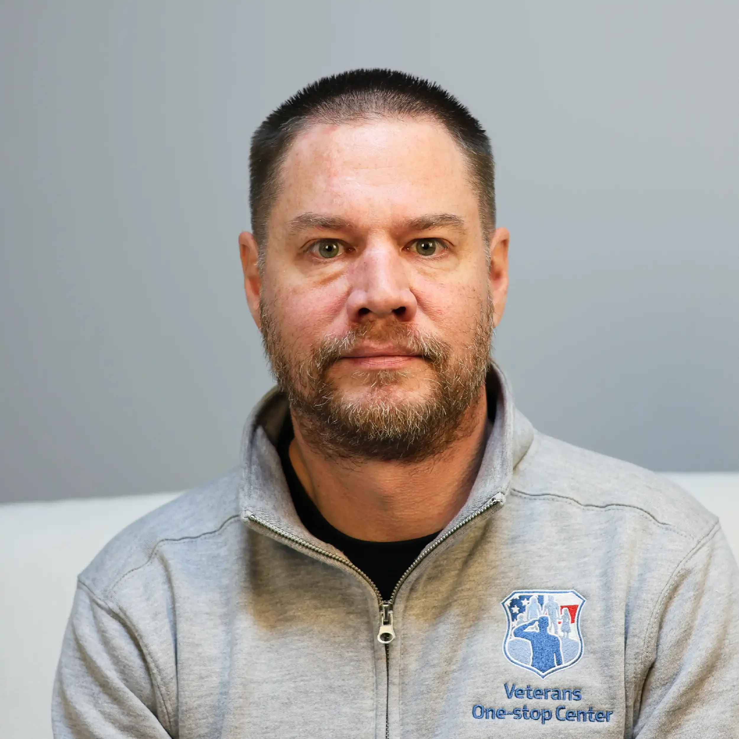A man with a beard and short dark hair wearing a gray jacket with the logo of Veterans One-Stop Center on it, sitting against a plain gray wall.