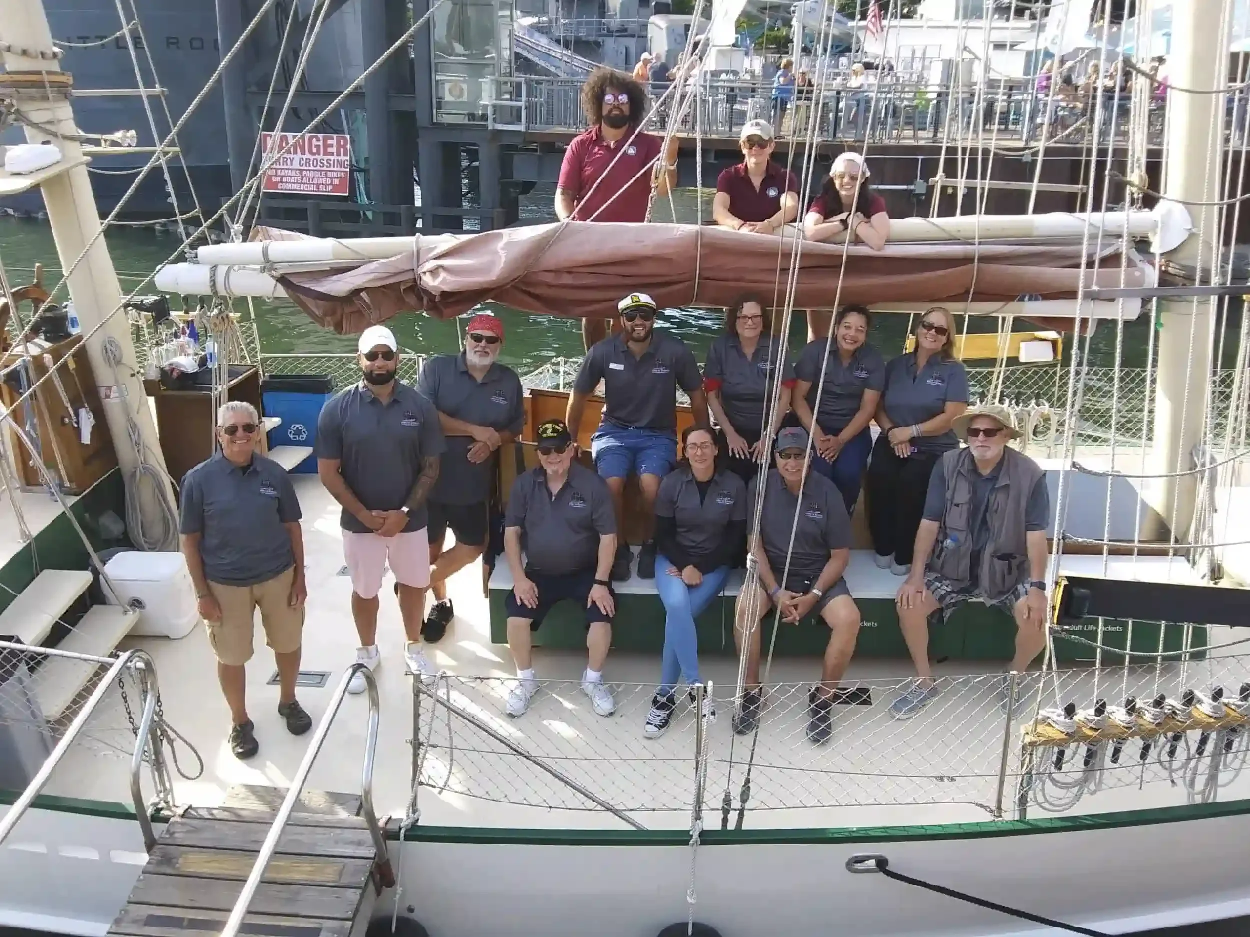 Group of people on a sailboat docked at a marina, some standing and some sitting, with the boat's sails and rigging visible.