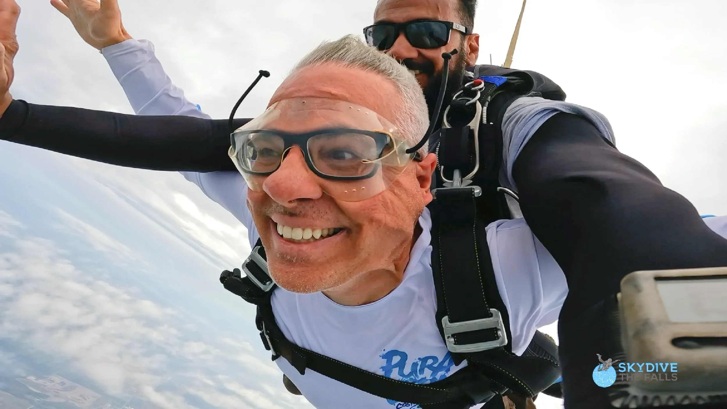 Two skydivers mid-air during a tandem jump, smiling and taking a selfie. The sky is partly cloudy, and they are wearing skydiving gear, including goggles and harnesses. One is an older man with gray hair, and the other is a younger man with sunglasses. The logo for 'Skydive The Falls' is visible in the bottom right corner.