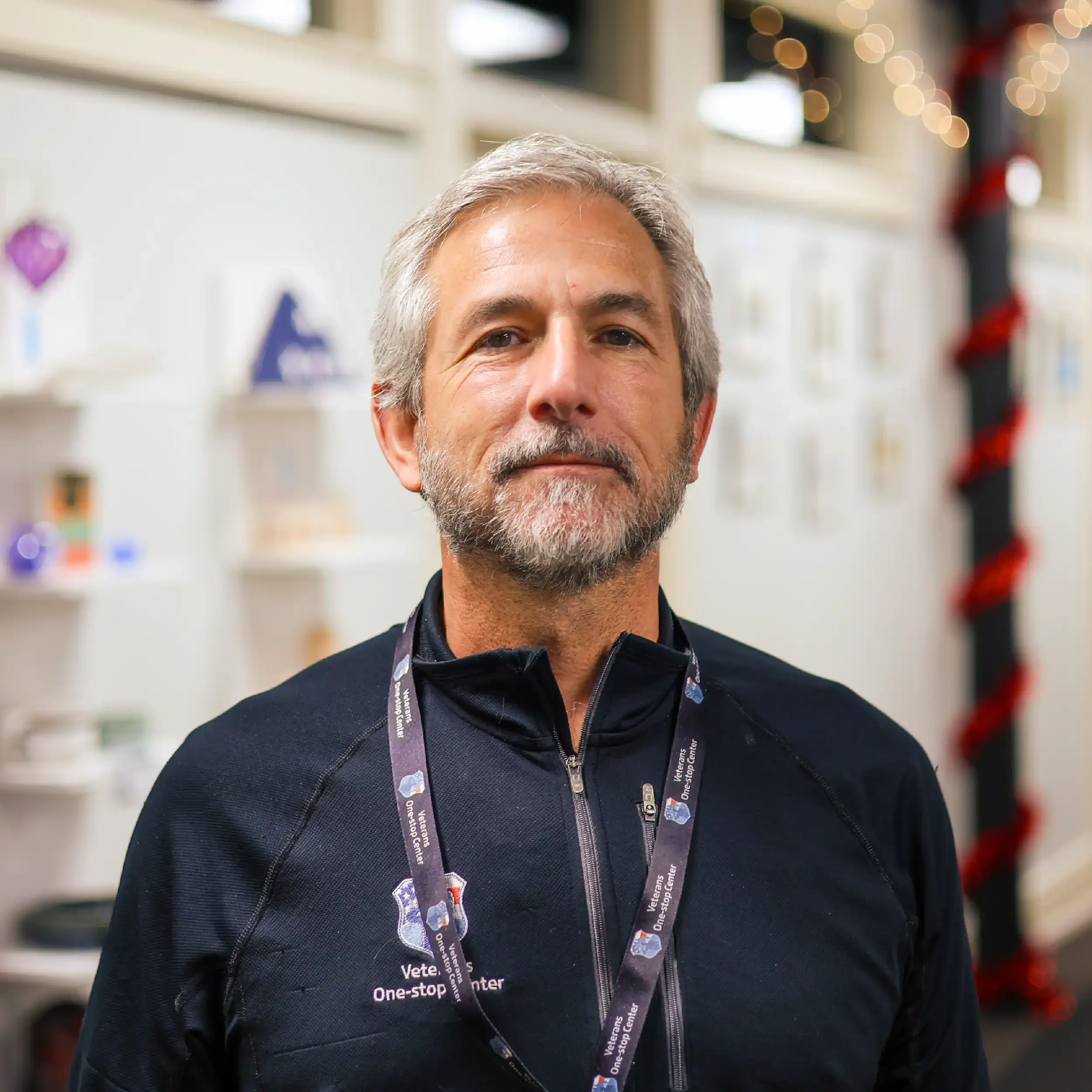 Portrait of a middle-aged man with gray hair and beard, wearing a black zip-up jacket and a lanyard, standing in a room with shelves and decorative items in the background.