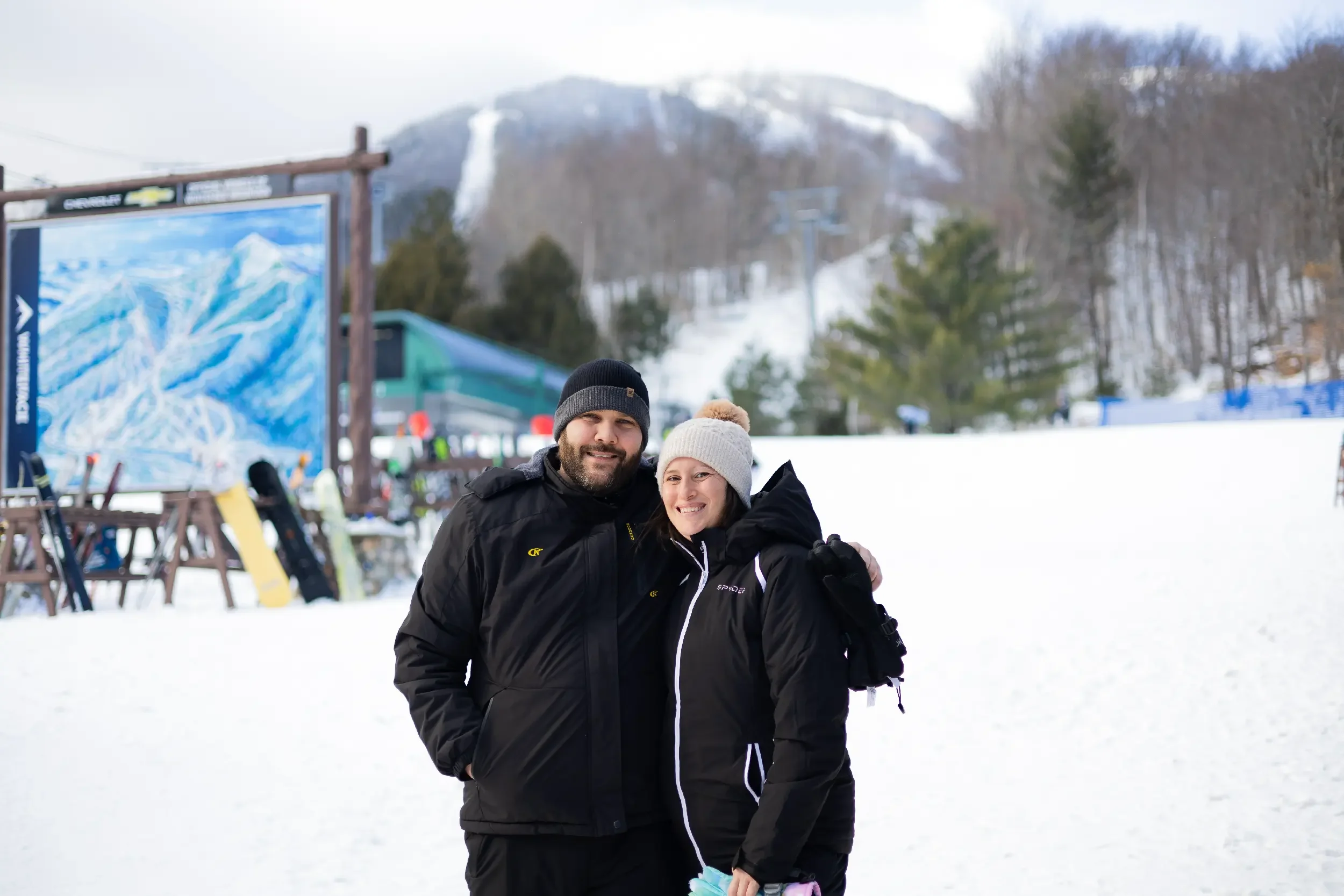 A smiling man and woman dressed in winter jackets and beanies, standing close together in a snowy ski resort, with ski equipment and a mountain in the background.