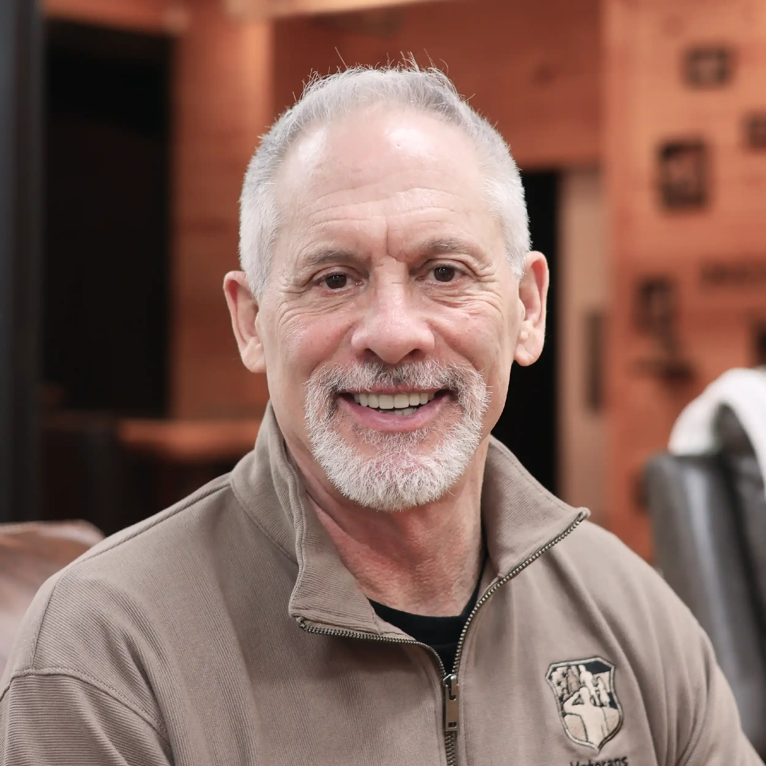 A smiling man with gray hair and a beard, wearing a beige jacket with a zipped-up collar and a badge on the chest, sitting indoors with wooden walls in the background.