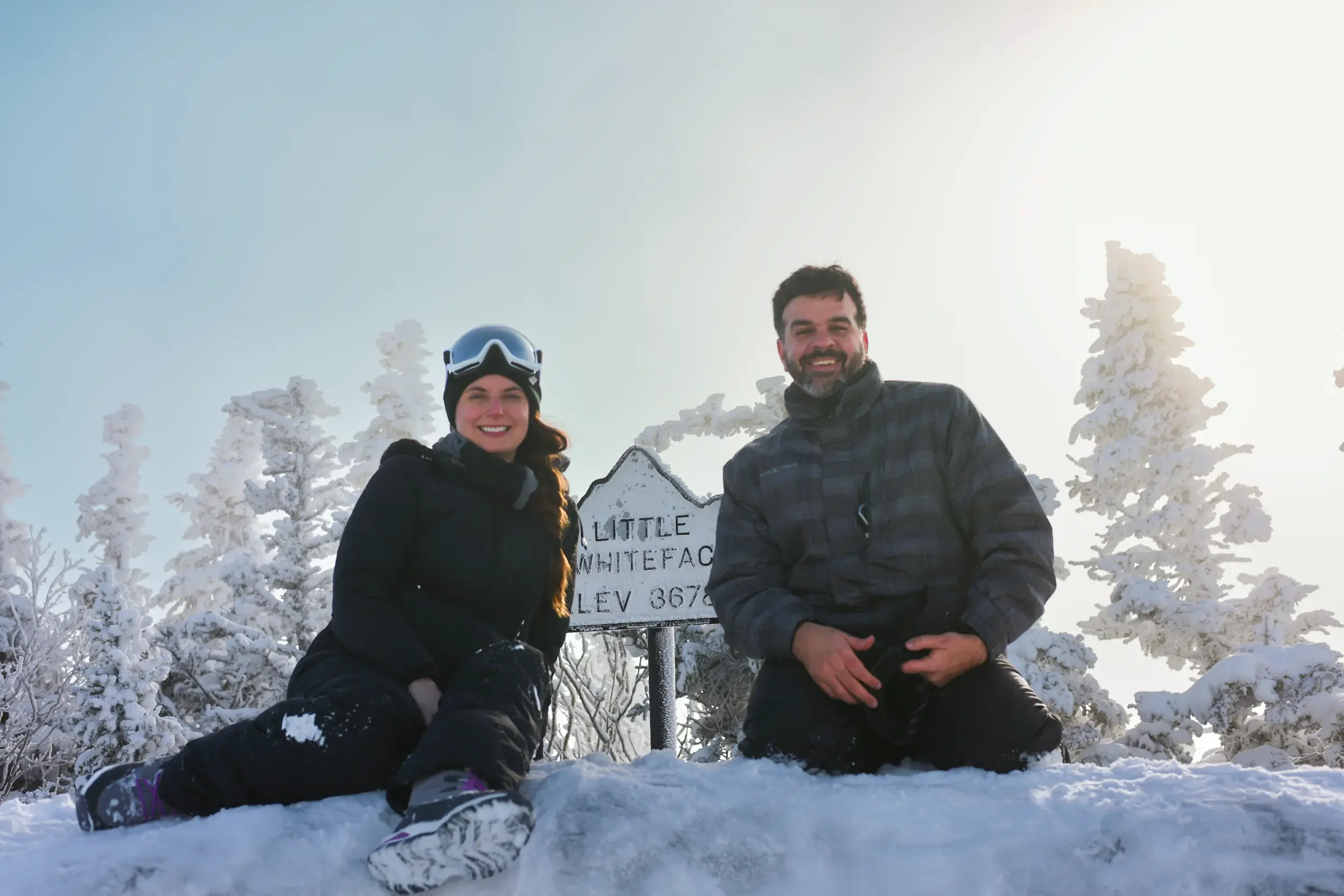 A man and woman kneeling in snow next to a sign that reads "LITTLE WHITEFACE ELEV 8670" on a snowy mountain with snow-covered trees and a bright sky in the background.