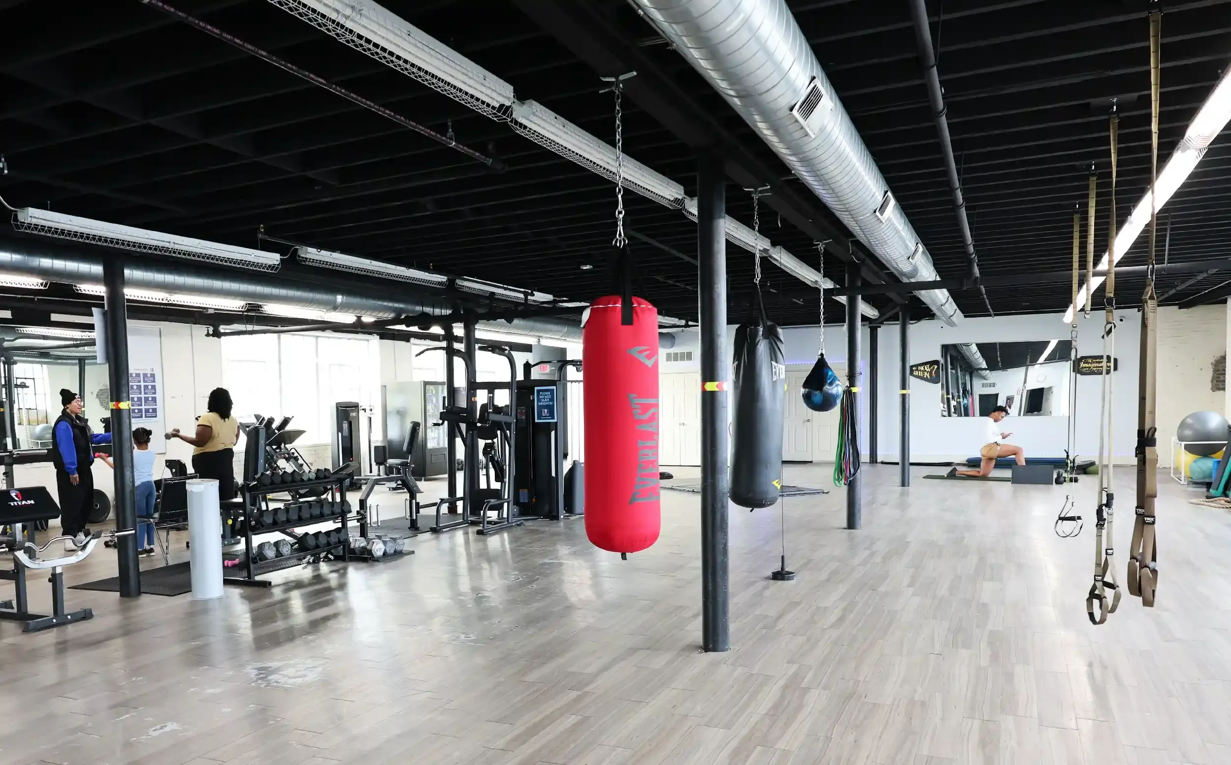 Large gym with various fitness equipment including punching bags, dumbbells, treadmills, and suspension trainers. There are people working out and socializing.