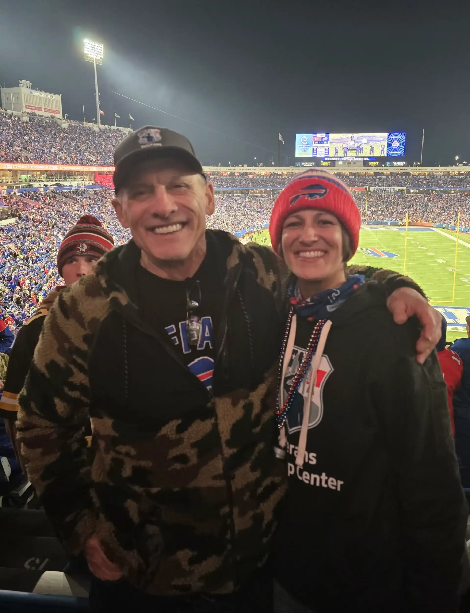 Two people smiling at a football game, one wearing a camouflage jacket and a baseball cap, the other wearing a red beanie and a black jacket, with a stadium filled with fans in the background.