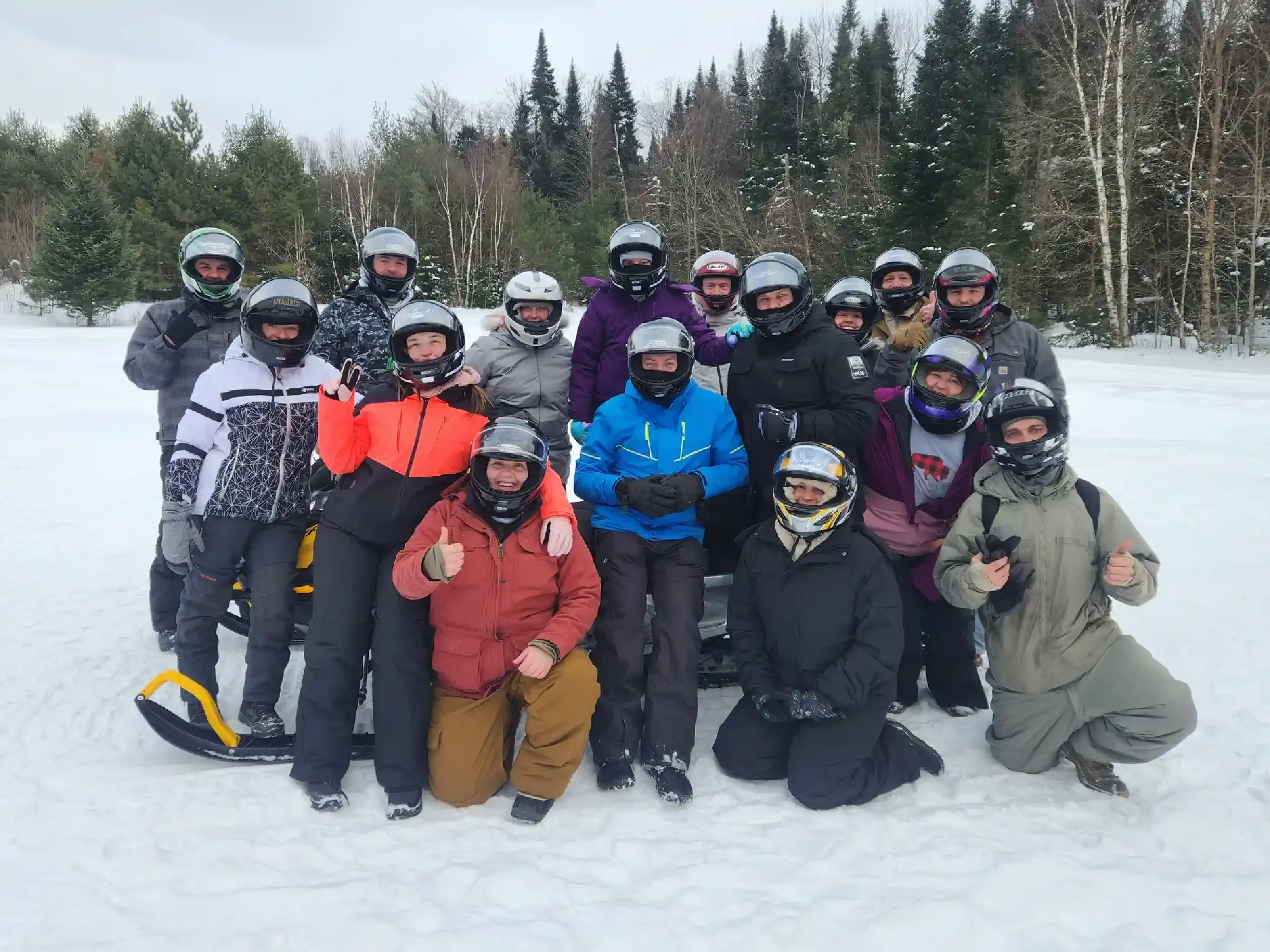 Group of people in winter gear and helmets posing on snowy ground with trees in background.