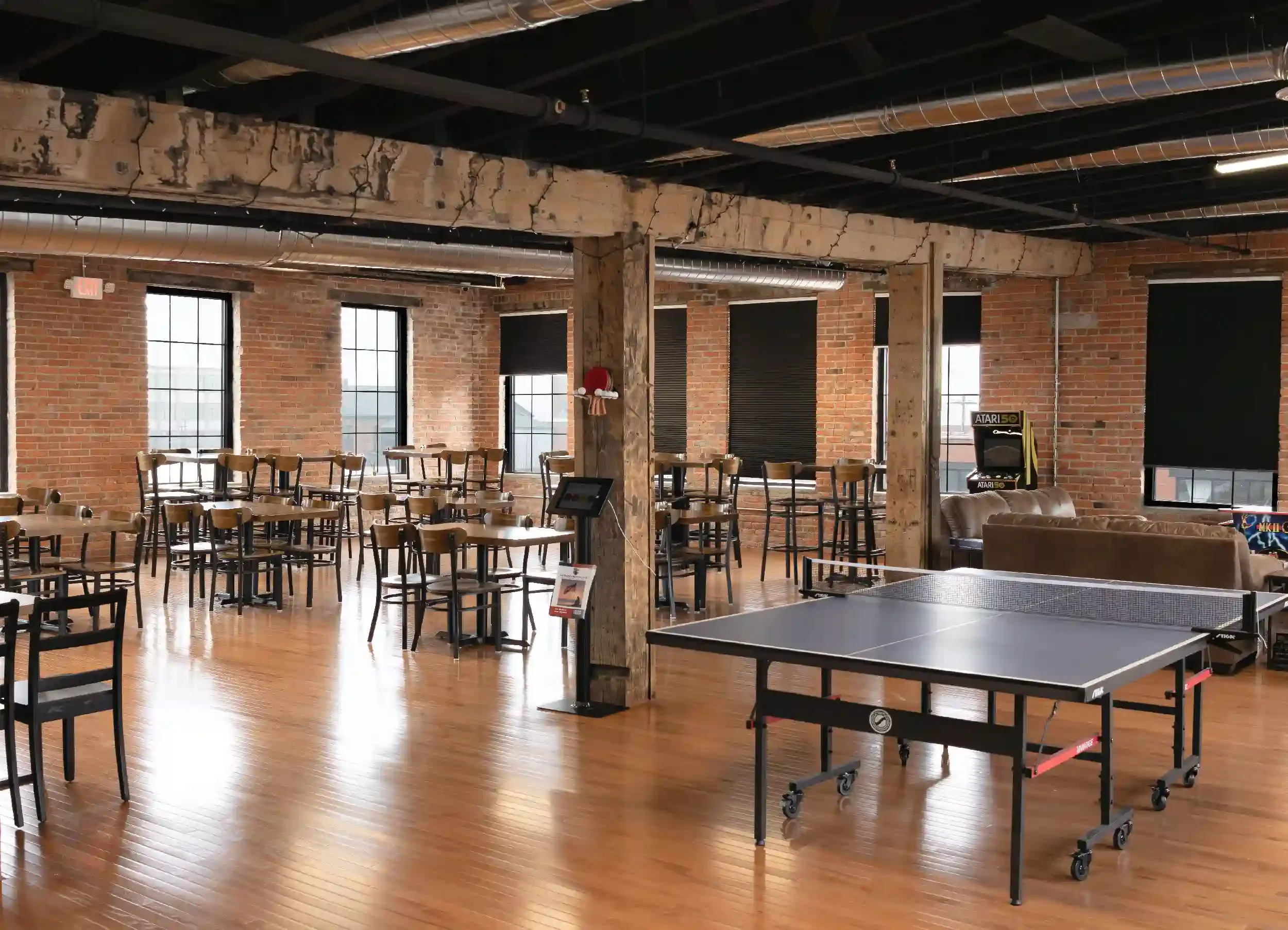 An empty recreational room with exposed brick walls and large windows, featuring a ping pong table, multiple high tables and chairs, a plush sofa, and arcade machines.