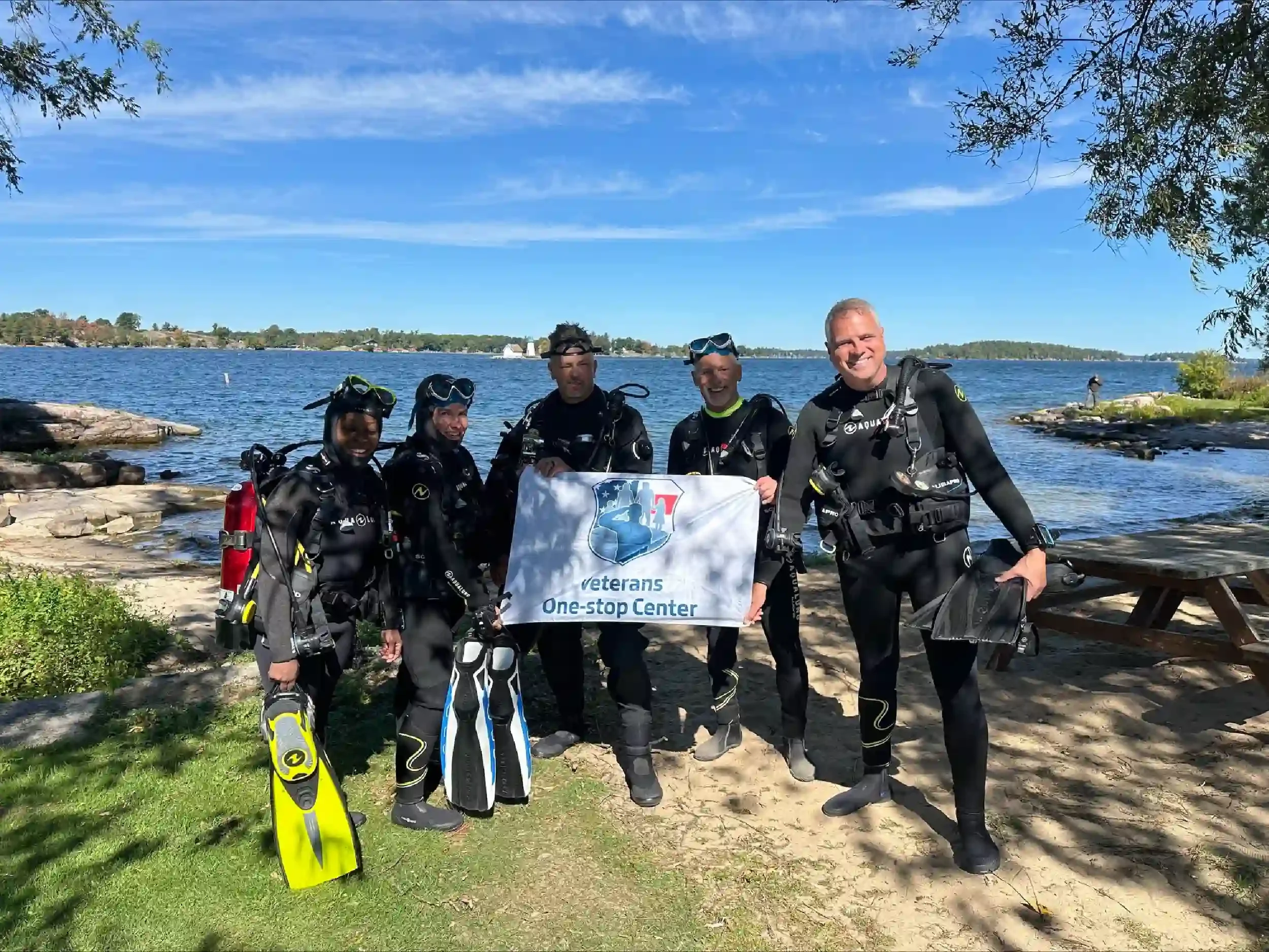 Group of five divers standing on a sandy and grassy shoreline holding a Veterans One-Stop Center banner, with a lake, trees, and a blue sky in the background.