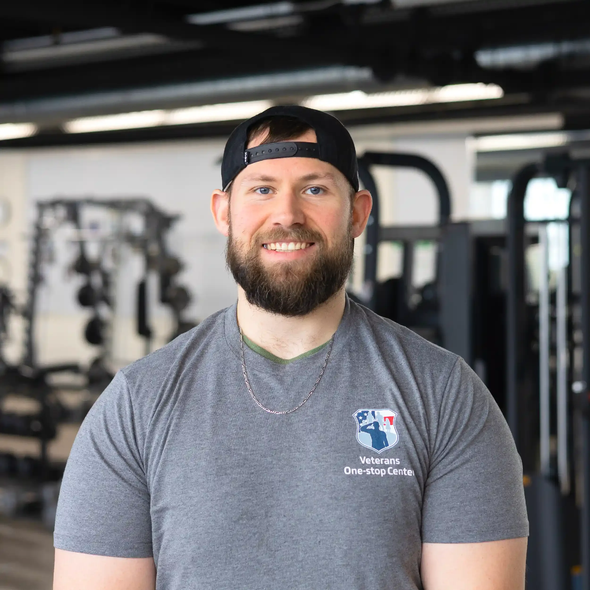 A smiling man with a beard and a black cap backwards, wearing a gray T-shirt with a 'Veterans One-stop Center' logo, standing in a gym with workout equipment in the background.