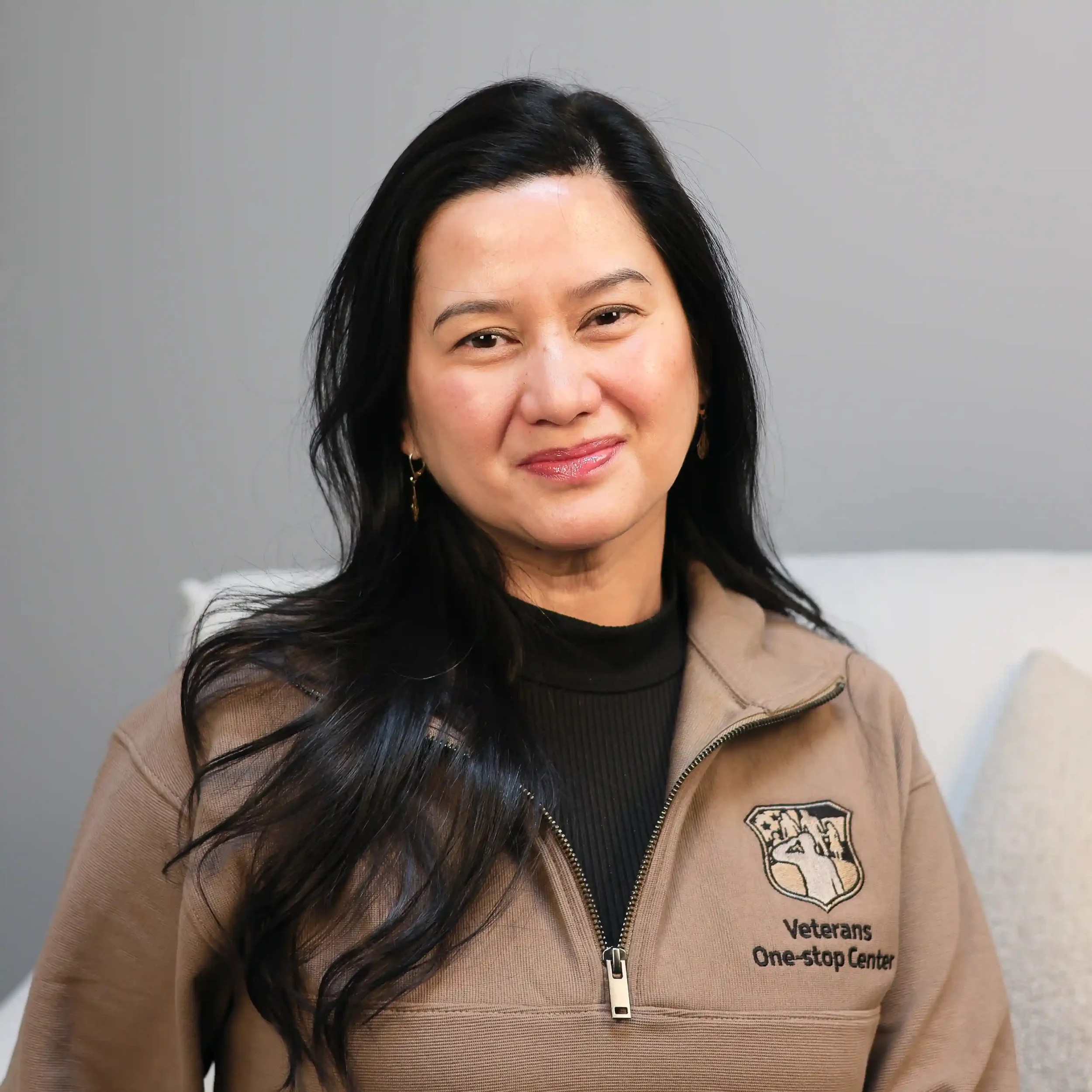 A woman with long black hair wearing a beige jacket with a Veterans One-Stop Center logo, sitting on a chair, smiling at the camera.