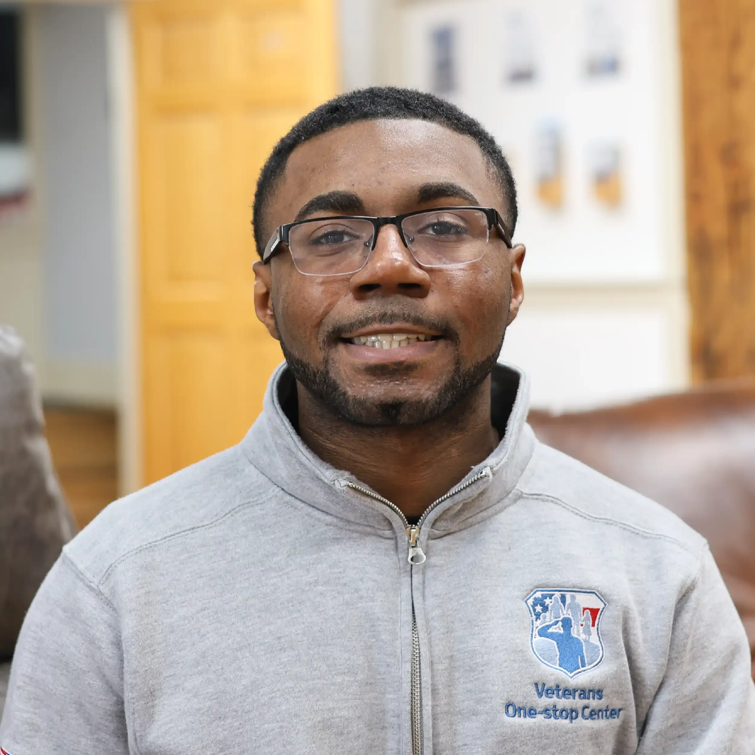 A man wearing glasses and a gray jacket with the Veterans One-Stop Center logo, sitting indoors with a blurred background of wooden cabinets and wall decorations.
