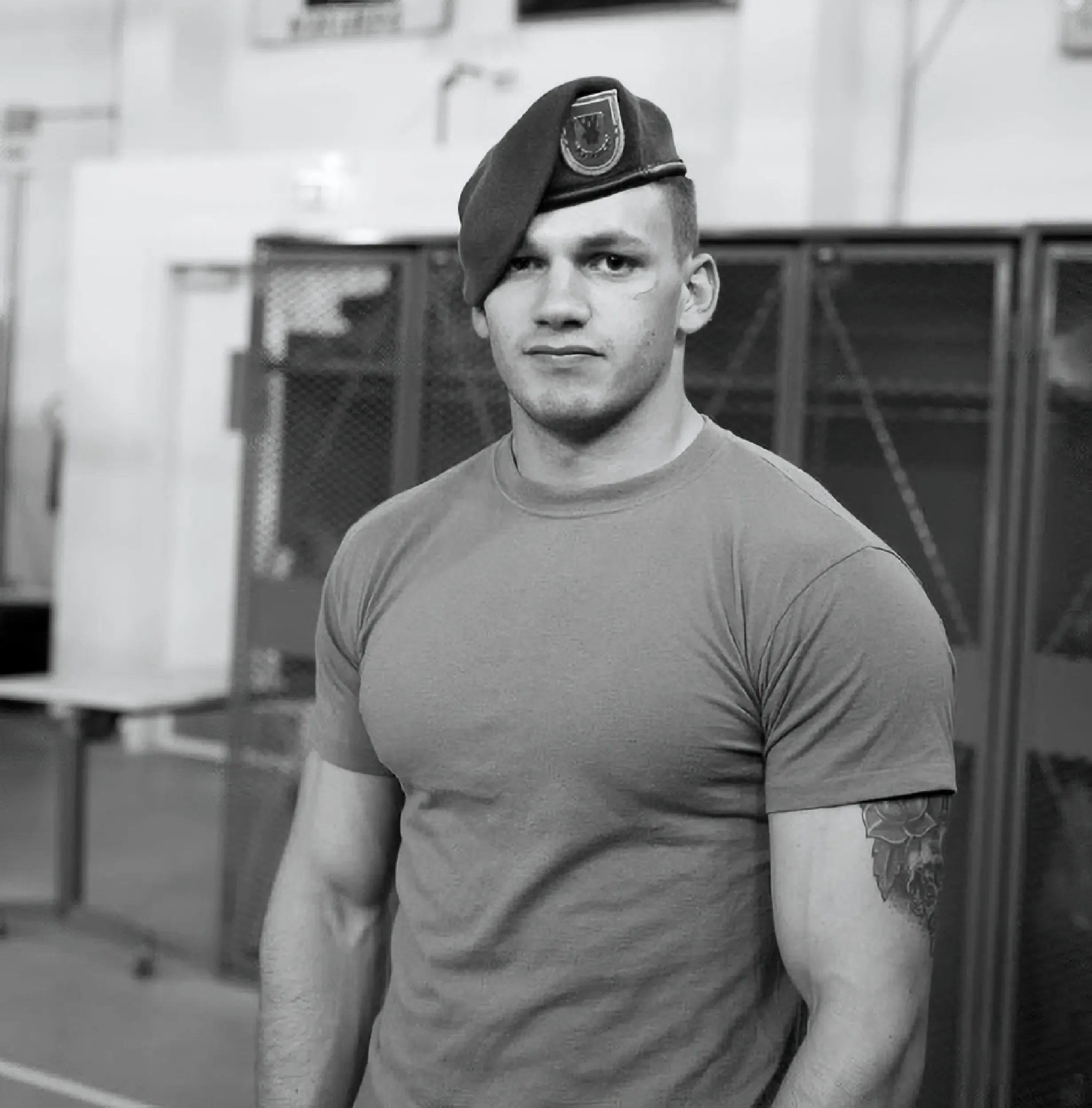 A young man wearing a military uniform beret with a badge, standing indoors in front of lockers, wearing a fitted t-shirt, with a tattoo on his left arm, looking at the camera.