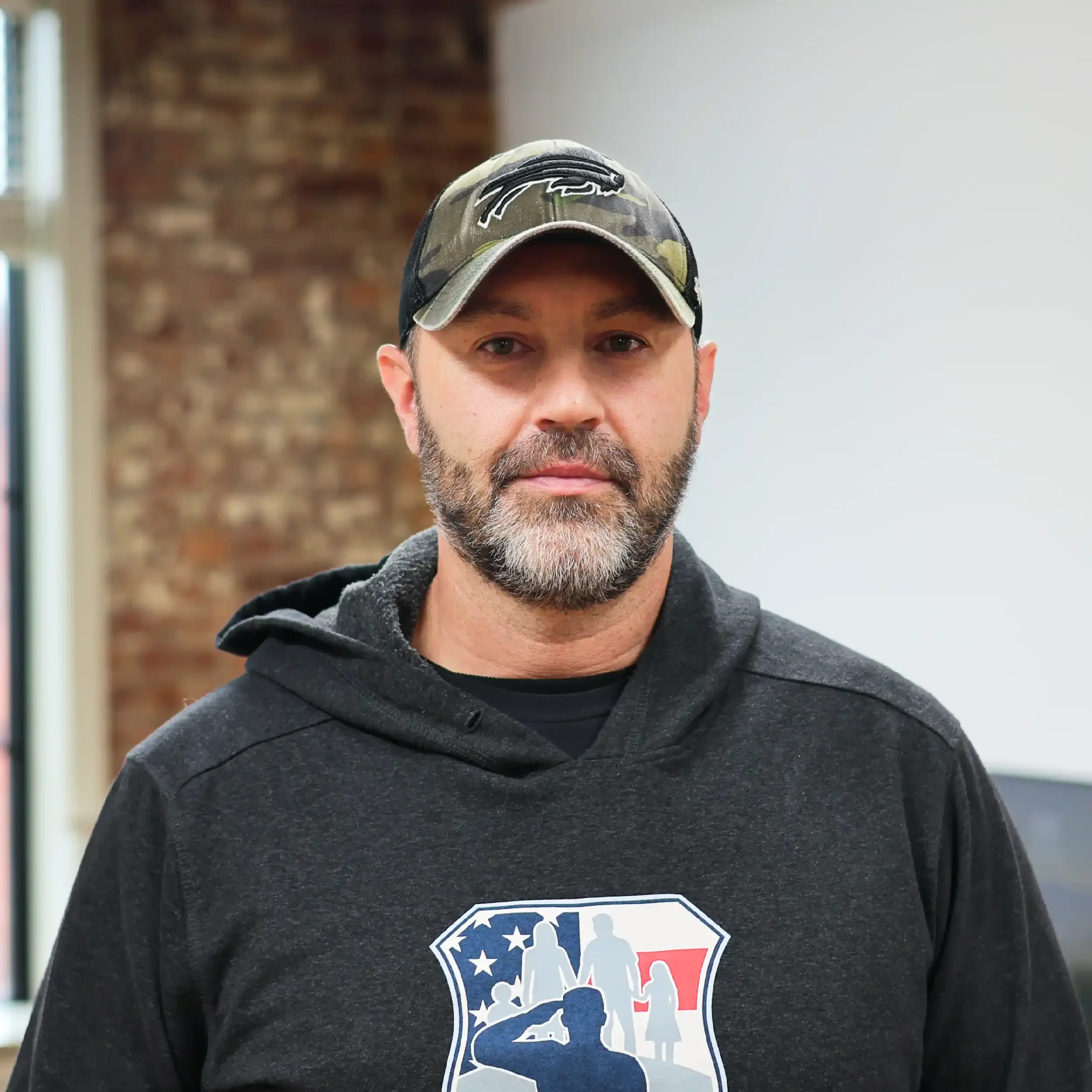 A man with a beard wearing a camouflage baseball cap and a black hoodie with a patriotic graphic.
