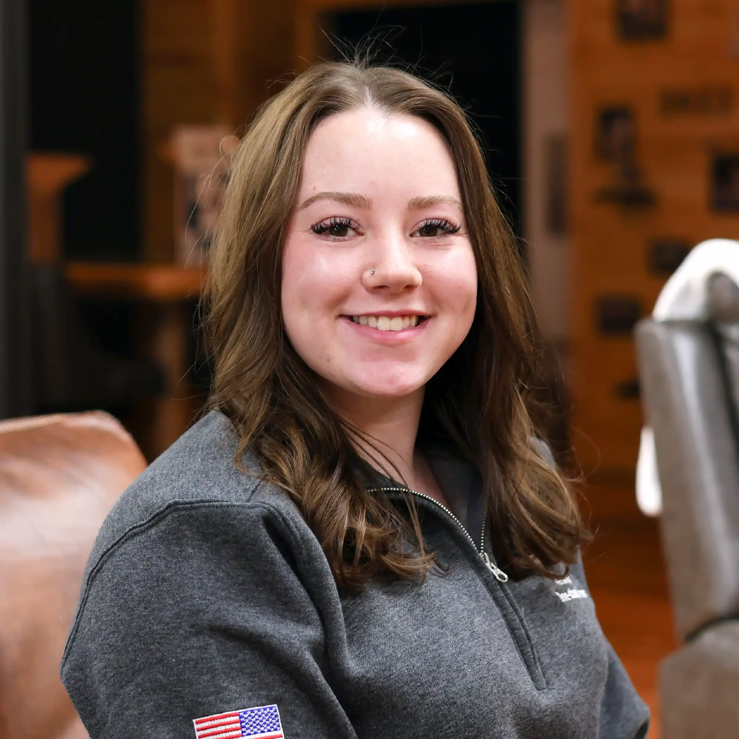 A young woman with wavy brown hair smiling at the camera, wearing a gray zip-up jacket with an American flag patch on the sleeve, sitting in a room with wooden walls and framed pictures in the background.