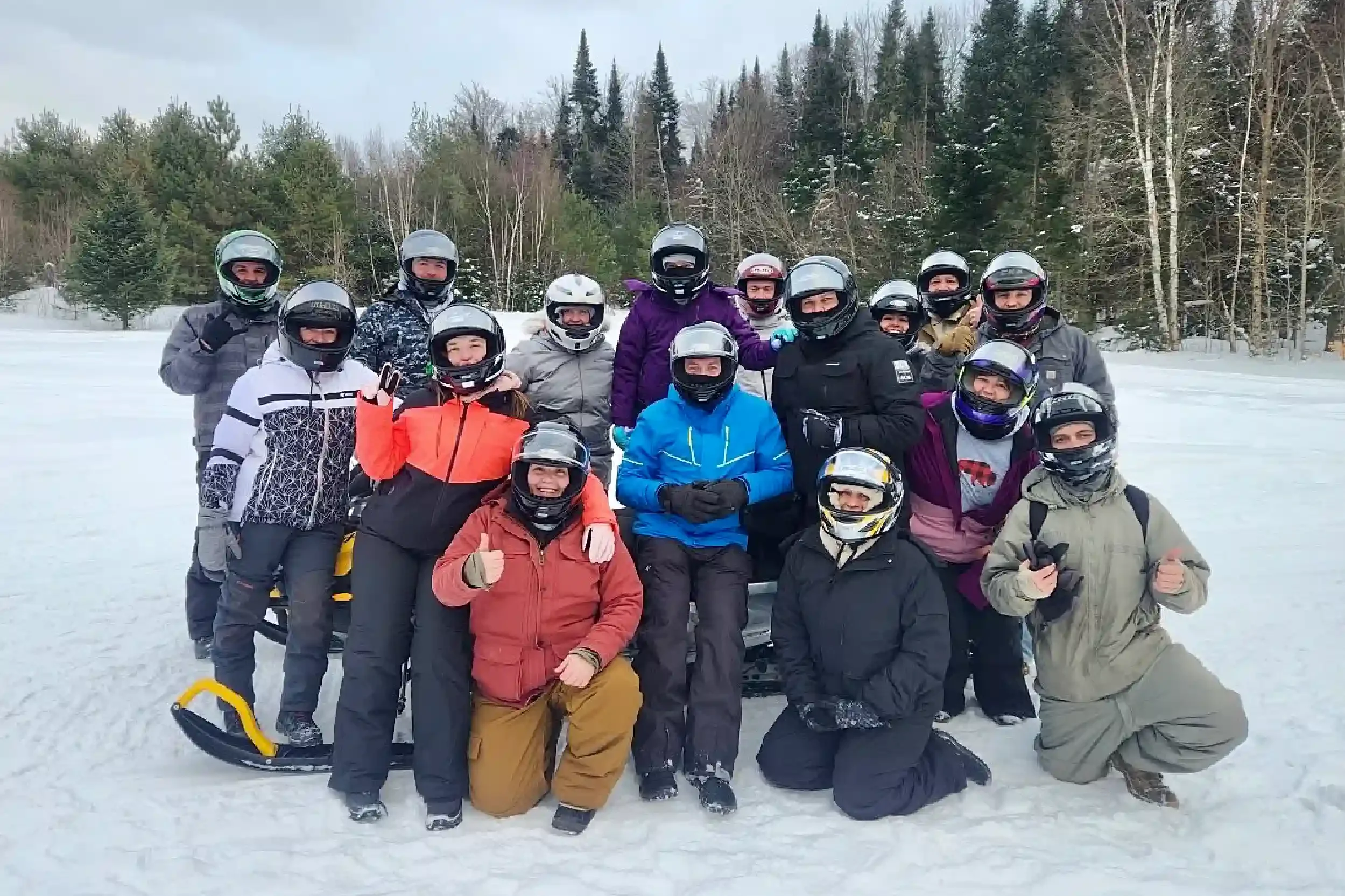 Group of people in winter clothing and helmets, posing outdoors on snow, with trees and a snowy landscape in the background.