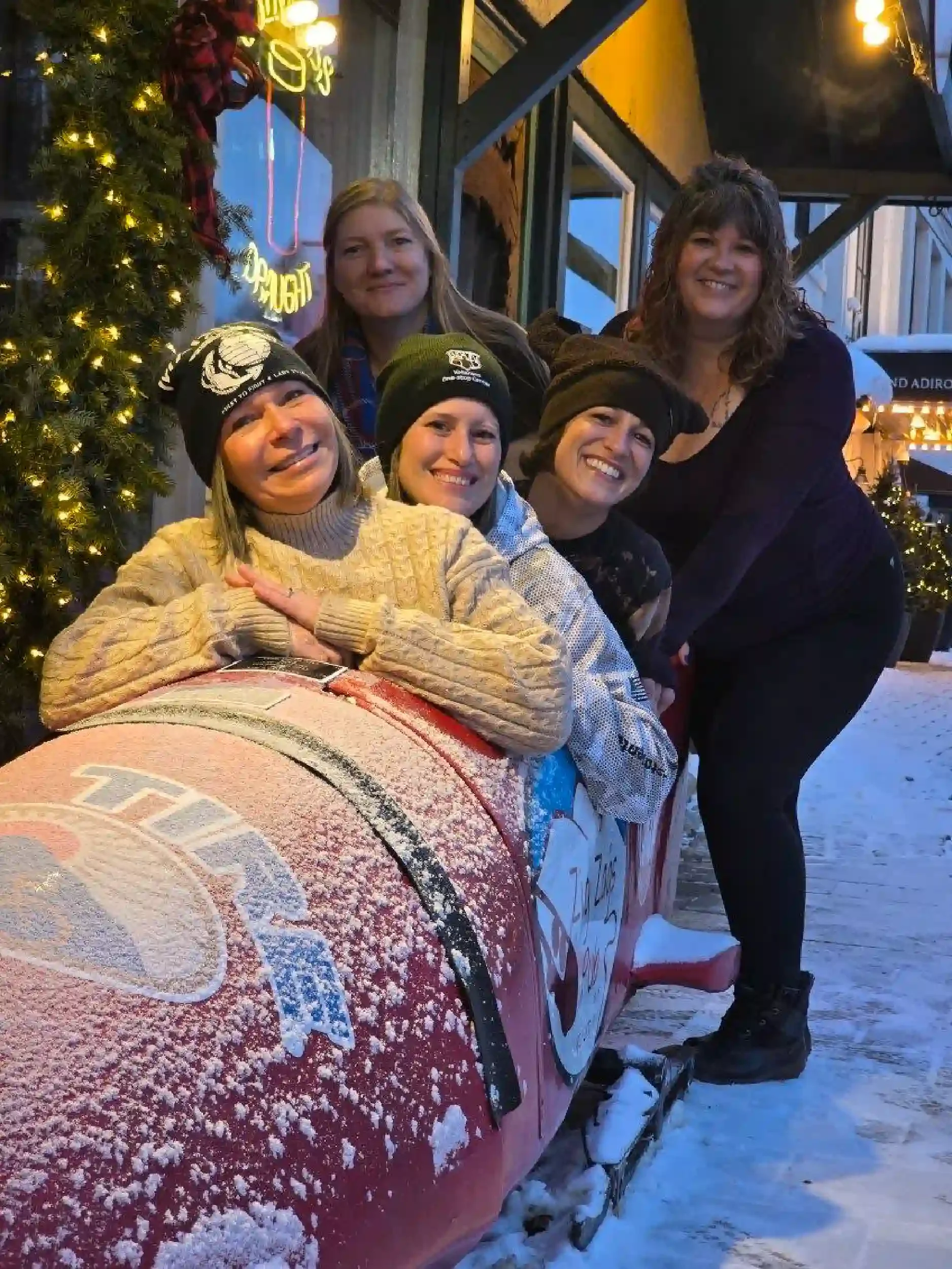 Four women and one girl gathered outside a restaurant or cafe during winter, with snow covering a red snowmobile in front of them. The women are smiling, some wearing black beanies, and the girl is wearing a beige sweater and a black beanie. Christma